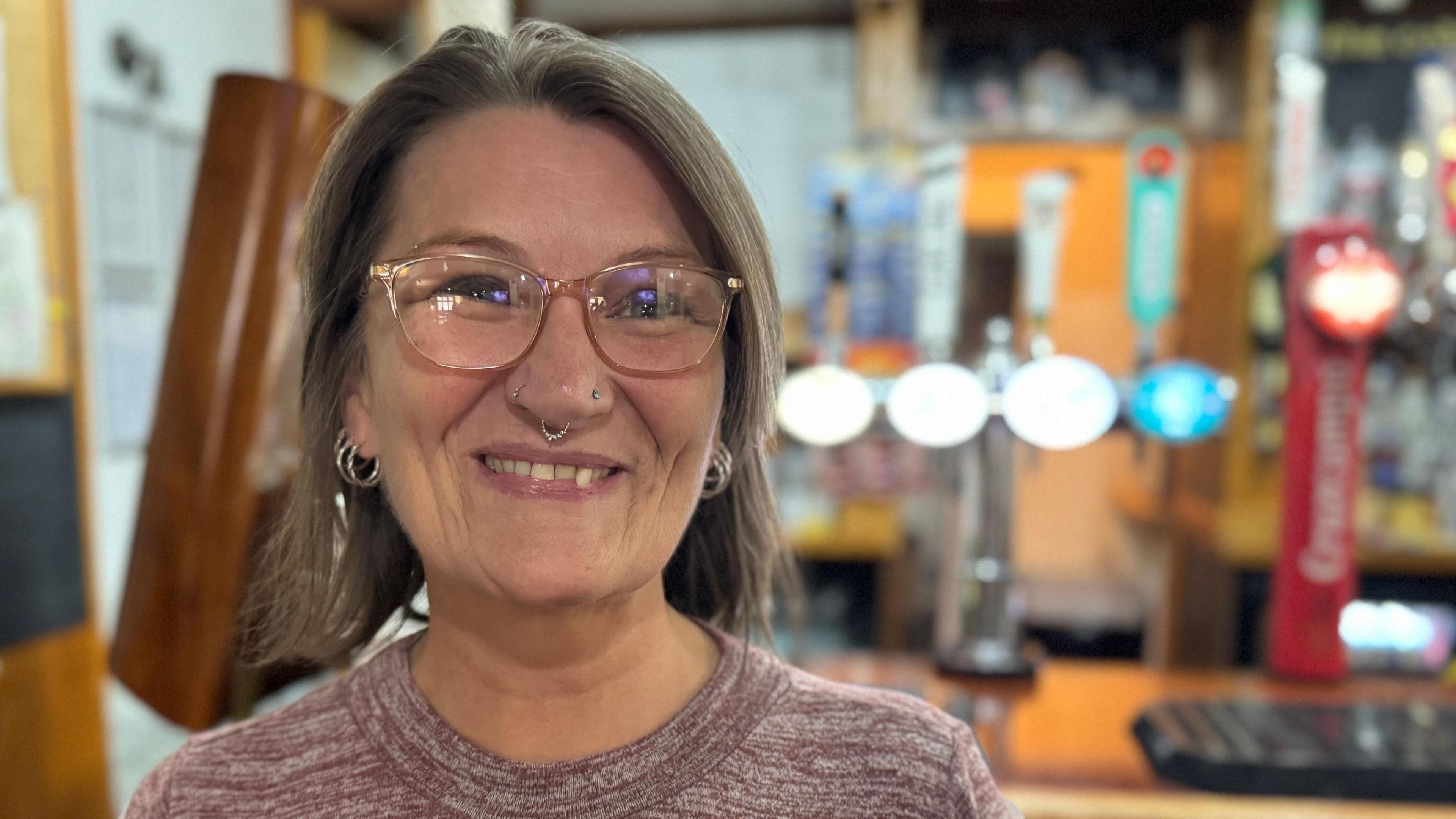 A woman with shoulder length hair in a round-neck top. She's wearing glasses and has earrings, a nose stud and nose ring. Standing in front of a pub bar.