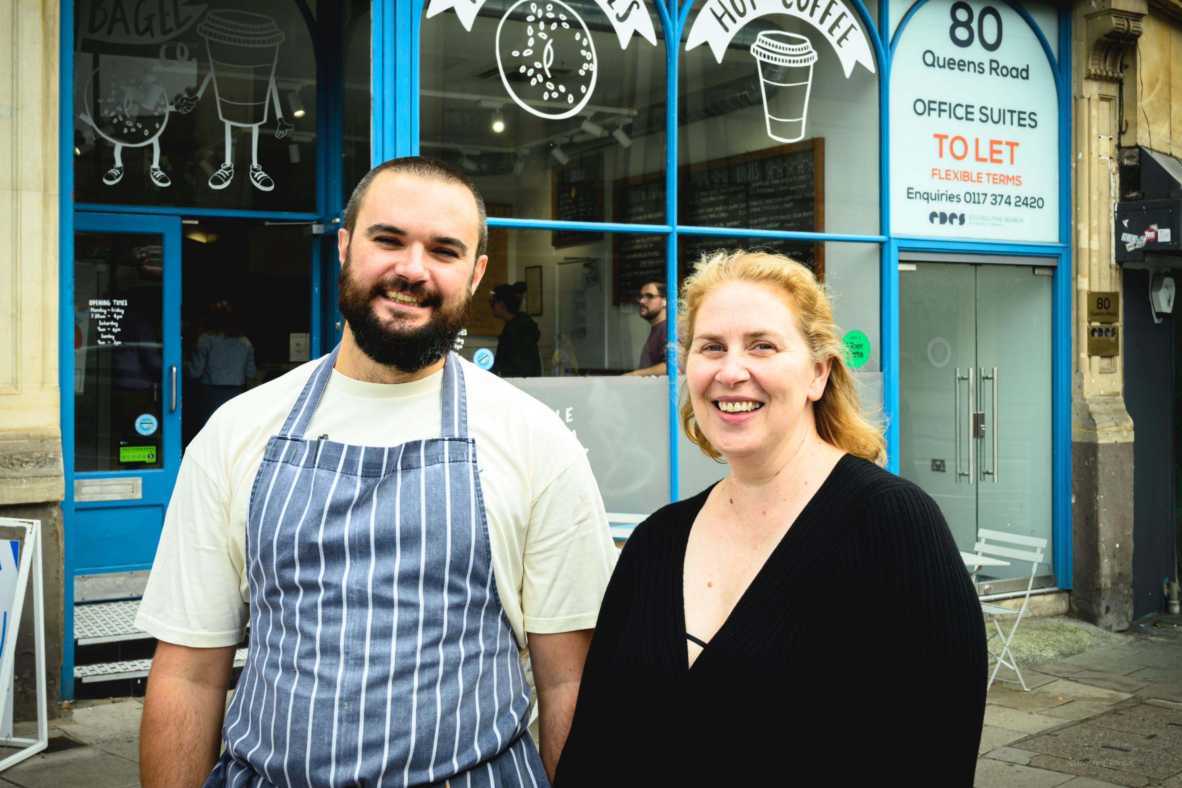 A couple stand outside a business premises in Bristol. On the left is a man with a white top and a blue and white vertically striped apron. He has a dark beard and short dark hair. The other person is a woman with fair shoulder-length hair and a black cardigan on