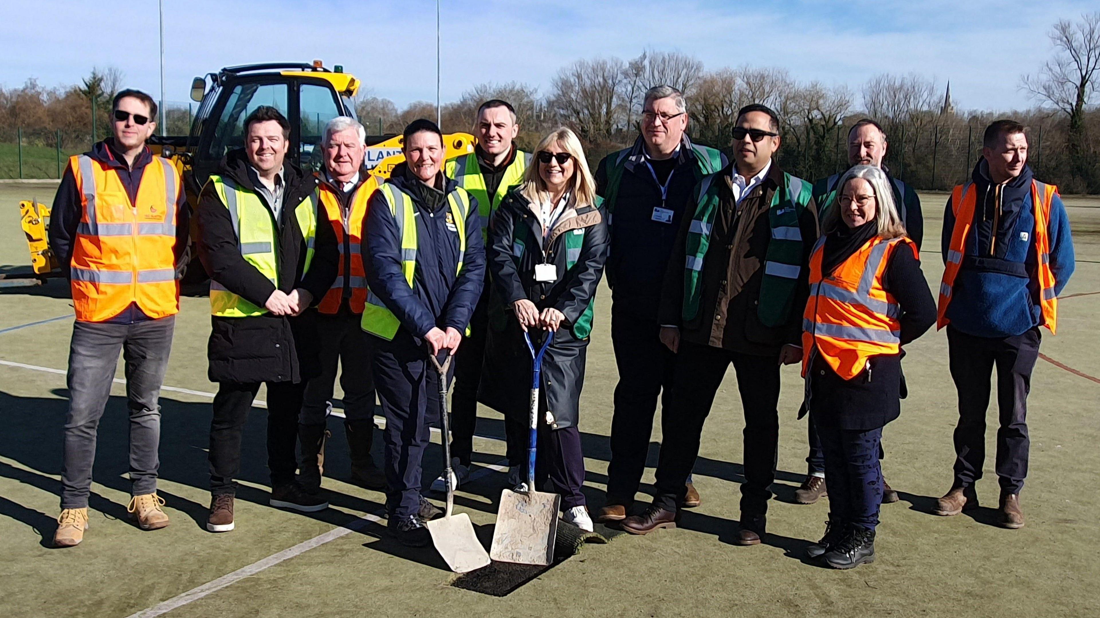 A group of people, some wearing orange hi-vis, surround a woman who is holding a blue-bladed spade.  A man alongside her also has a spade. The blads are in a cut-out section of the surface of a decayed artificial football pitch. There is a yellow digger behind the group and a house in background.