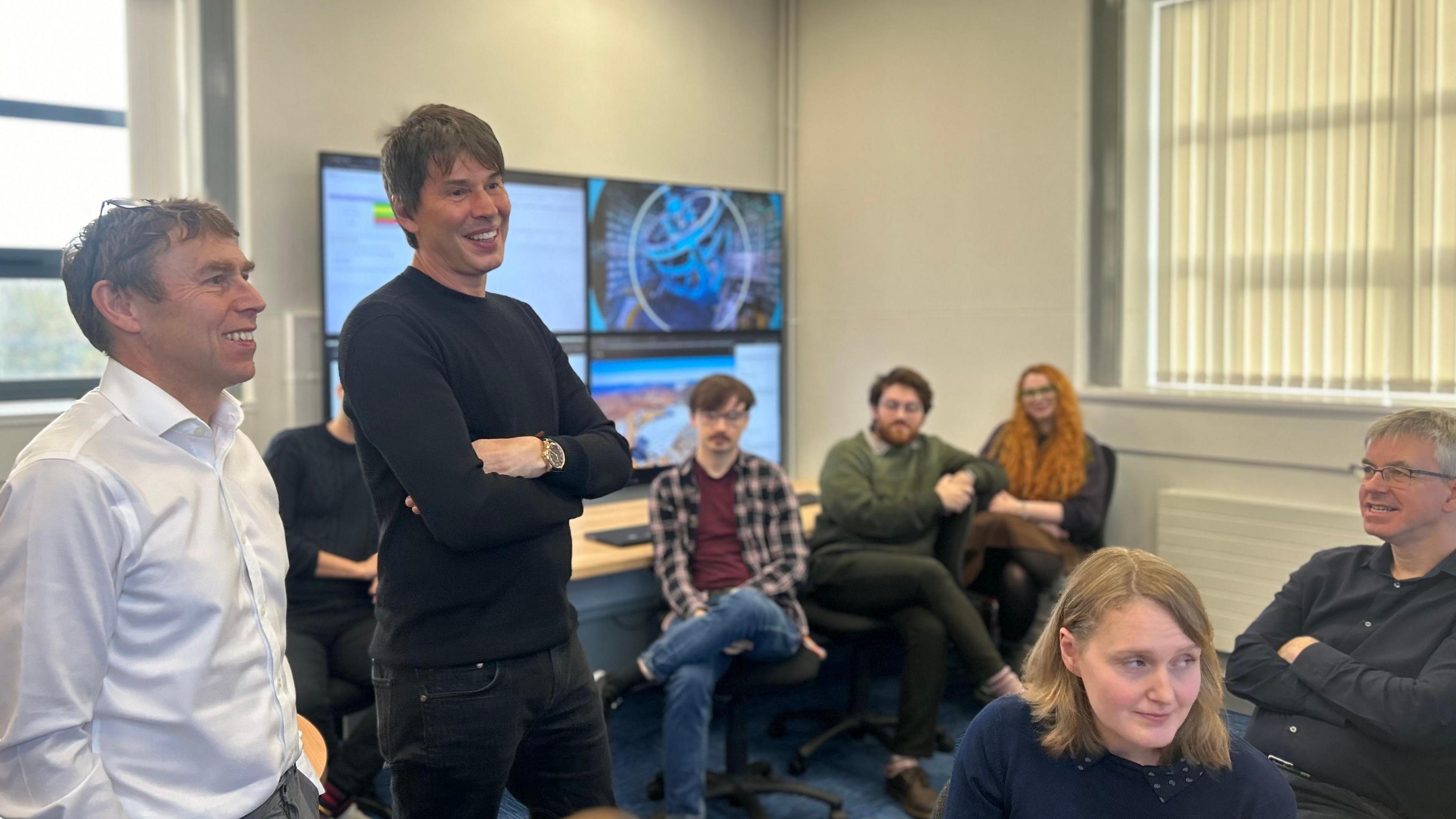 Students and teachers gathered around Prof Brian Cox as he speaks to them.