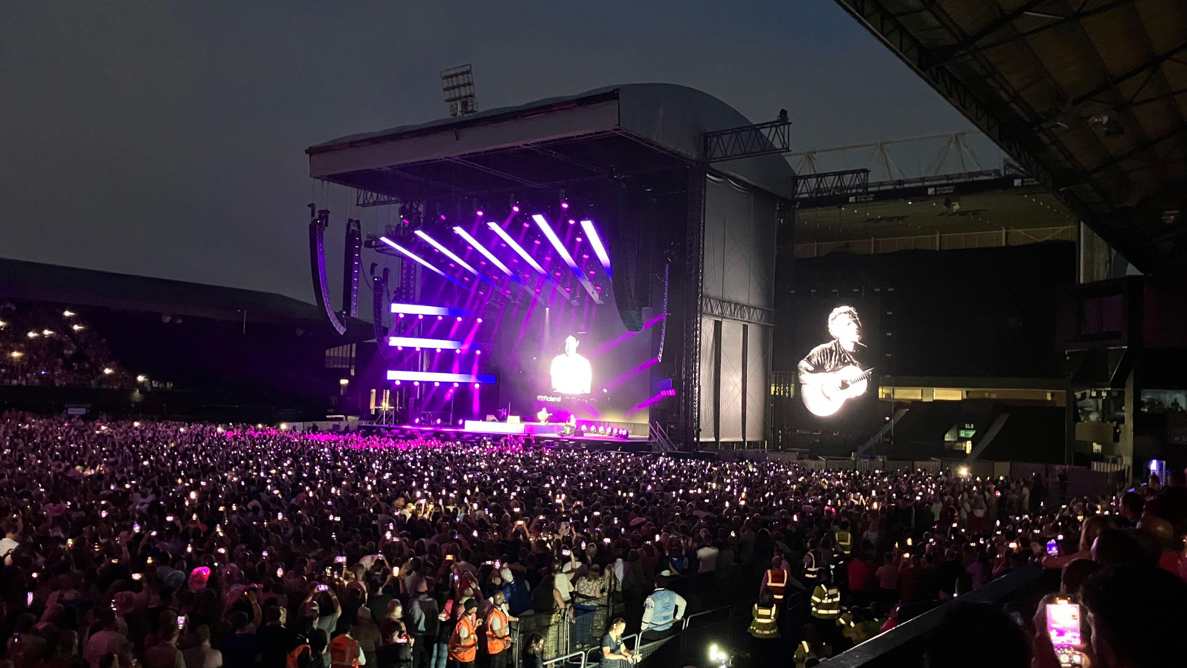 Ed Sheeran performs on stage in Portman Road. It is dark and the stage is lit up. Thousands of fans watch on.