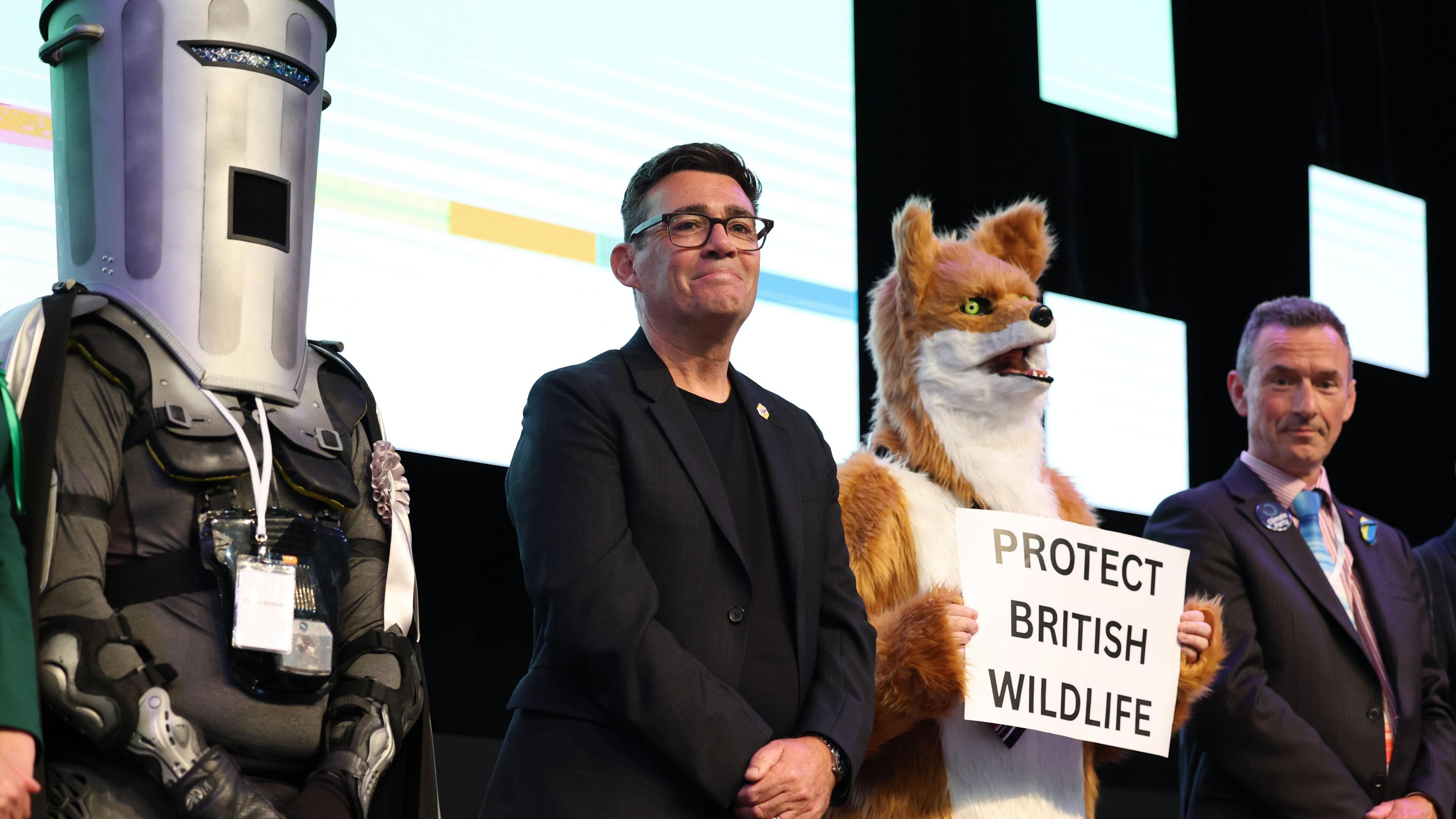 By-election count Makerfield. Andy Burnham stands between Count Binface with bin on his face and Insdependent ~Rob Pownall dressed as a fox.