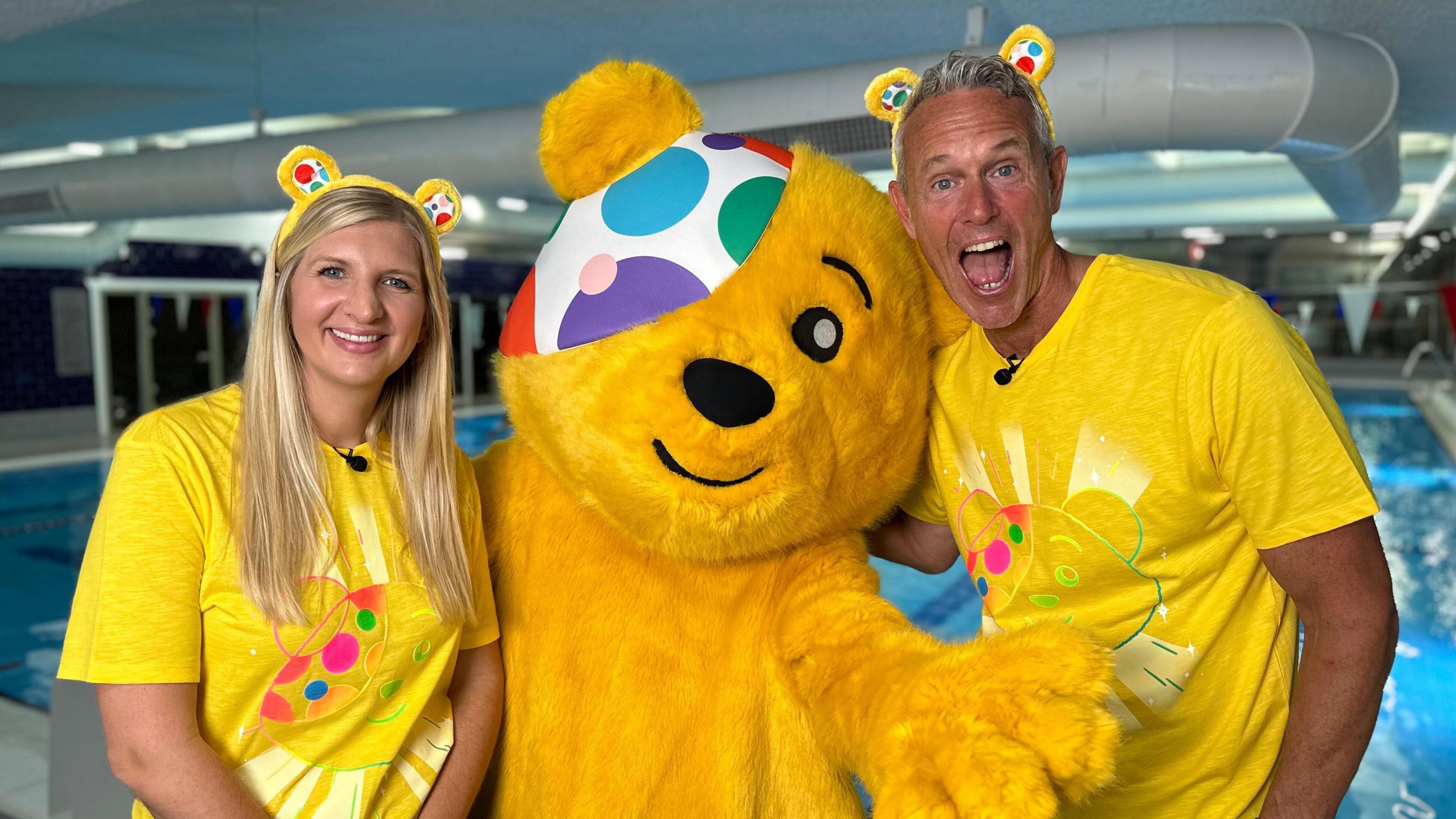 A giant Pudsey Bear stands next to an indoor swimming pool with Rebecca Adlington on his right and Mark Foster on his left. Rebecca is wearing yellow Pudsey Bear ears and a yellow T-shirt featuring Pudsey's face. Mark is wearing the same. Pudsey's left arm is stretched out in front of him. Rebecca is smiling. Mark is pulling an excited face with his mouth wide open