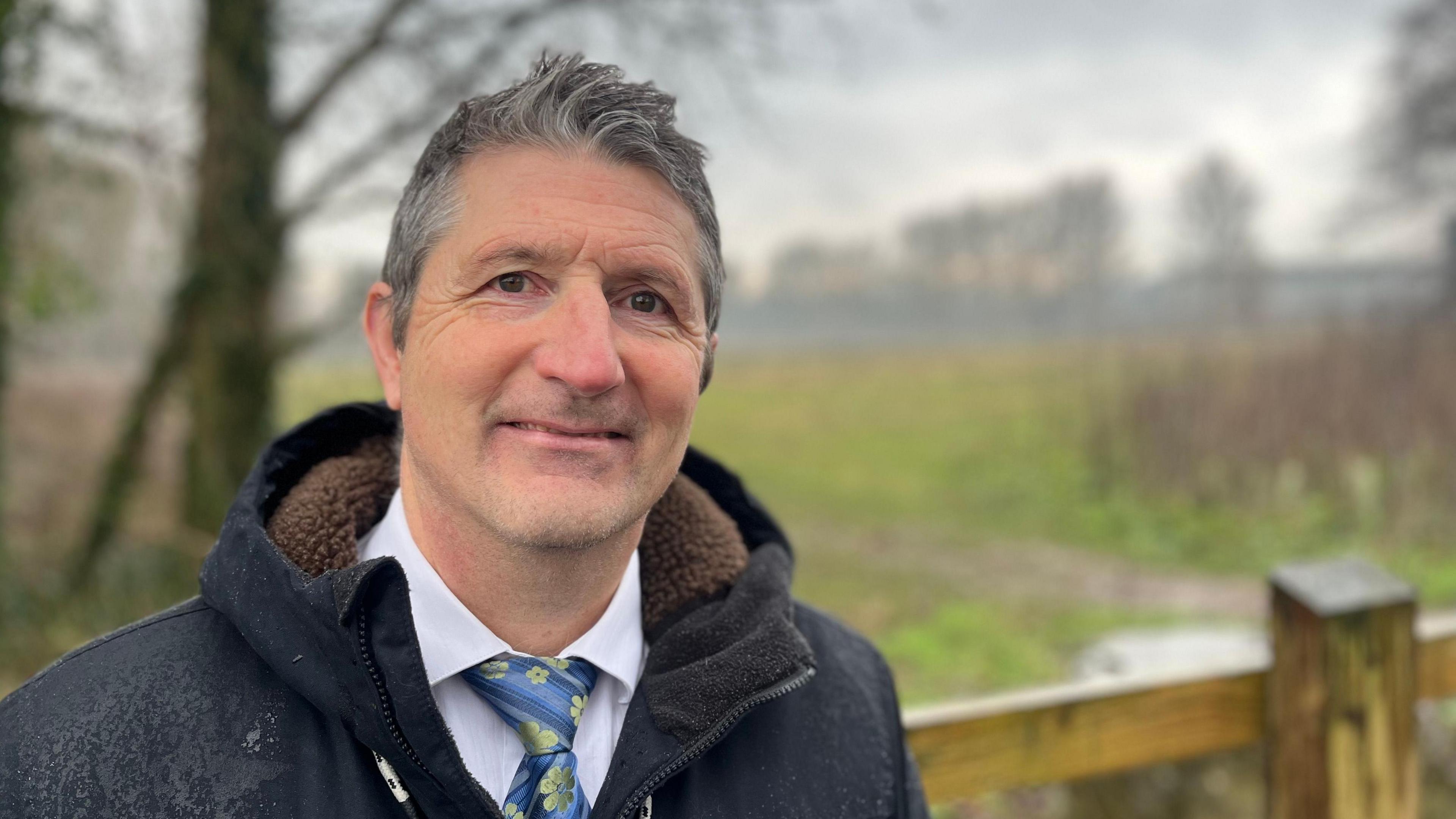 Green Party councillor Andy Ketchin wearing a white shirt and blue and yellow tie and a black coat, standing in front of the field in the Riverside Valley Park