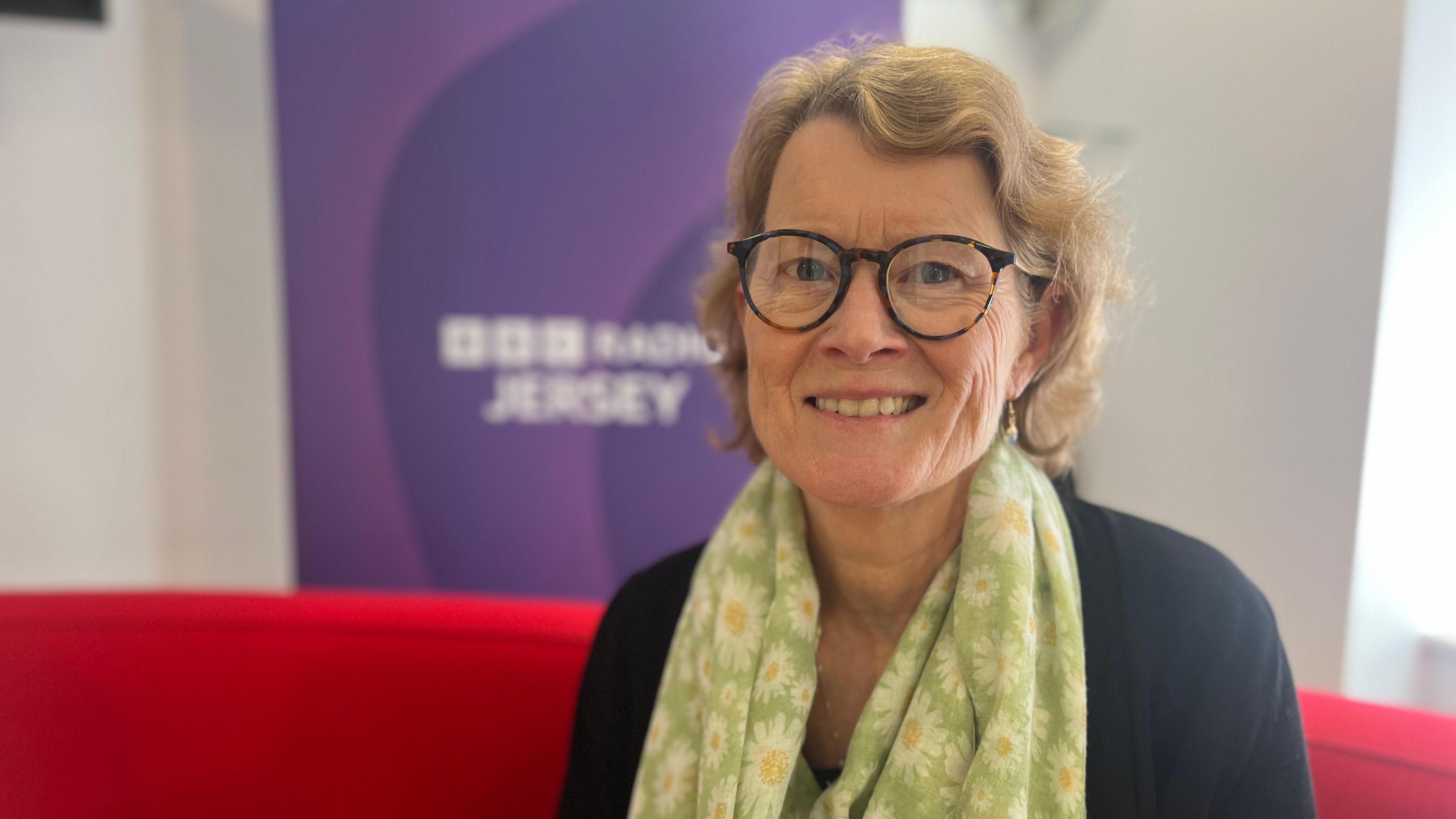 Caroline Spencer smiles at the camera as she sits on the red sofa with a BBC Radio Jersey banner behind her and she's wearing a scarf with a sunflower pattern on it