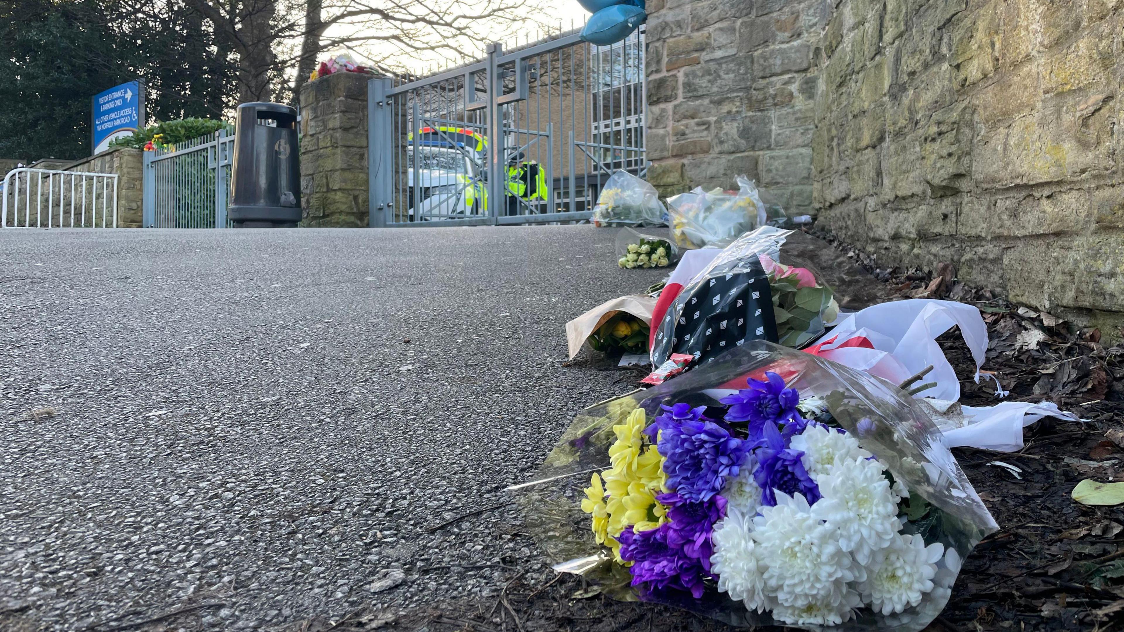 Floral tributes line the school's wall. A police car and officer can be seen behind the metal school gates. Blue balloons have been tied to the entrance on the right.