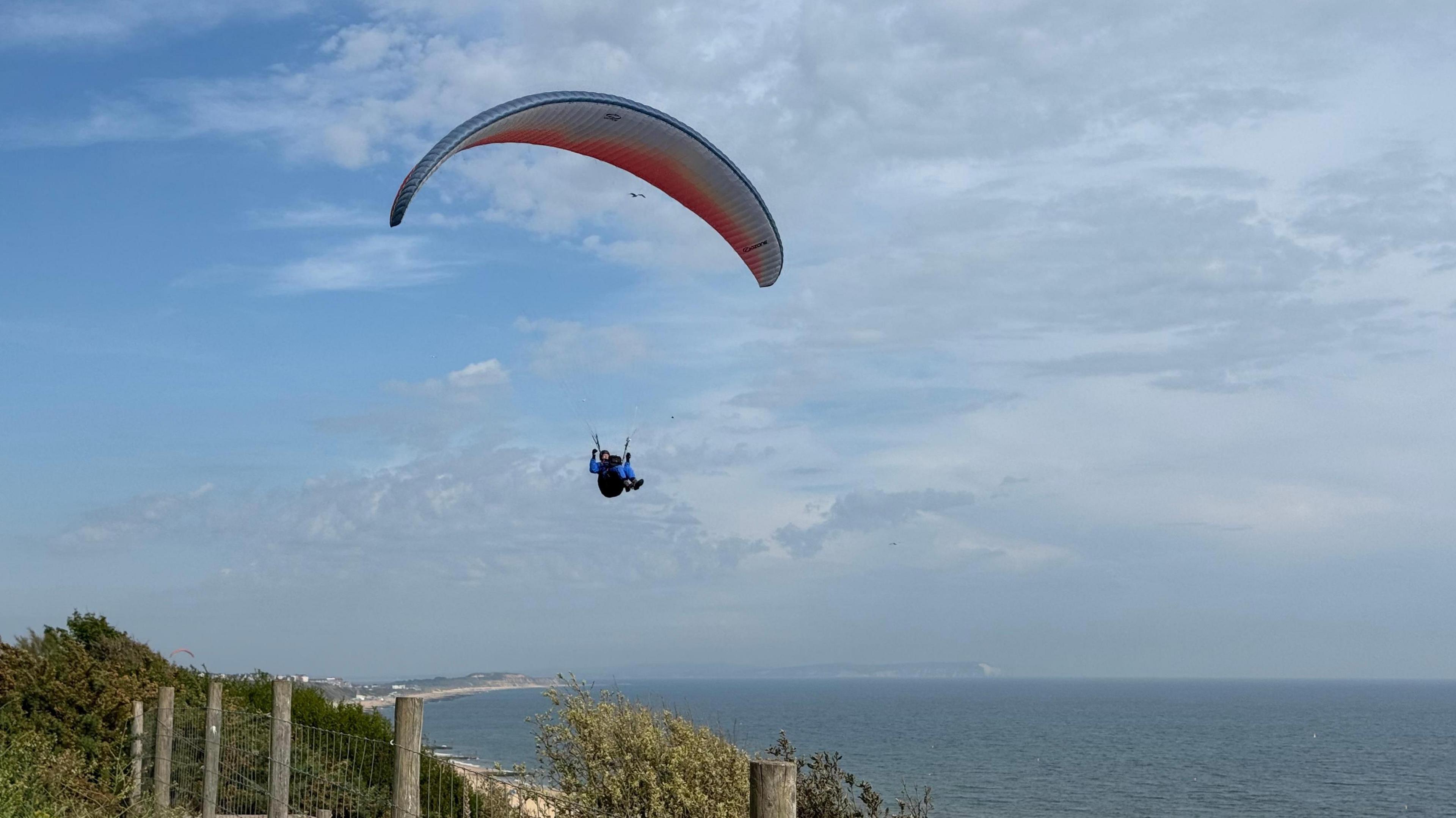 A paraglider floats over the coastline with the sea behind him and a beach trailing into the distance
