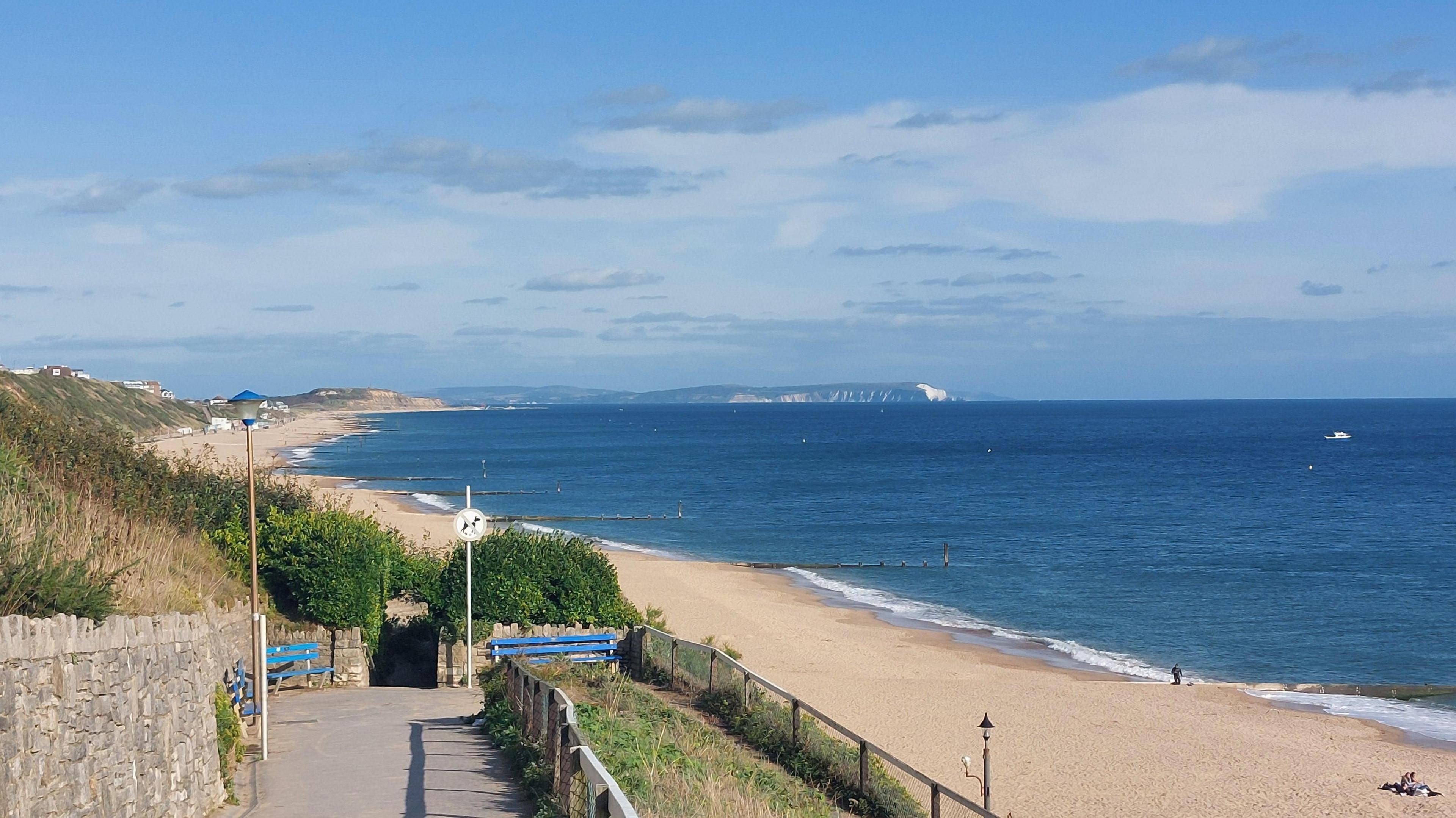 A long stretch of golden beach, lapped by sea waves, can be seen from the cliff above stretching towards the horizon, with blue skies above