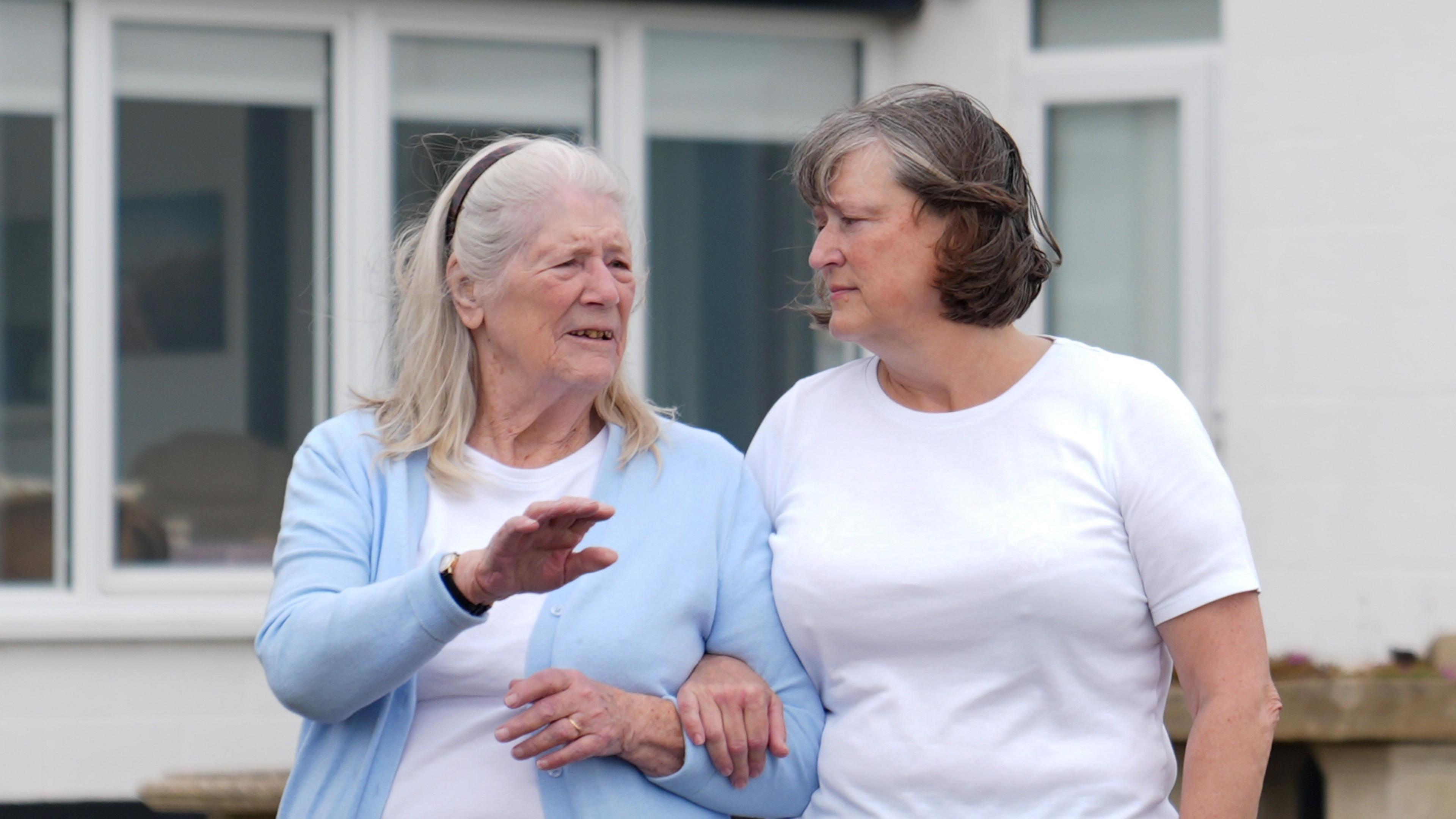 Jean Flick stands arm in arm with her daughter Frances outside her home. They are mid conversation. Frances has shoulder length grey hair with a headband on. She wears a light blue cardigan with a white top underneath. Frances has short brown and grey hair, she wears a white T-shirt.