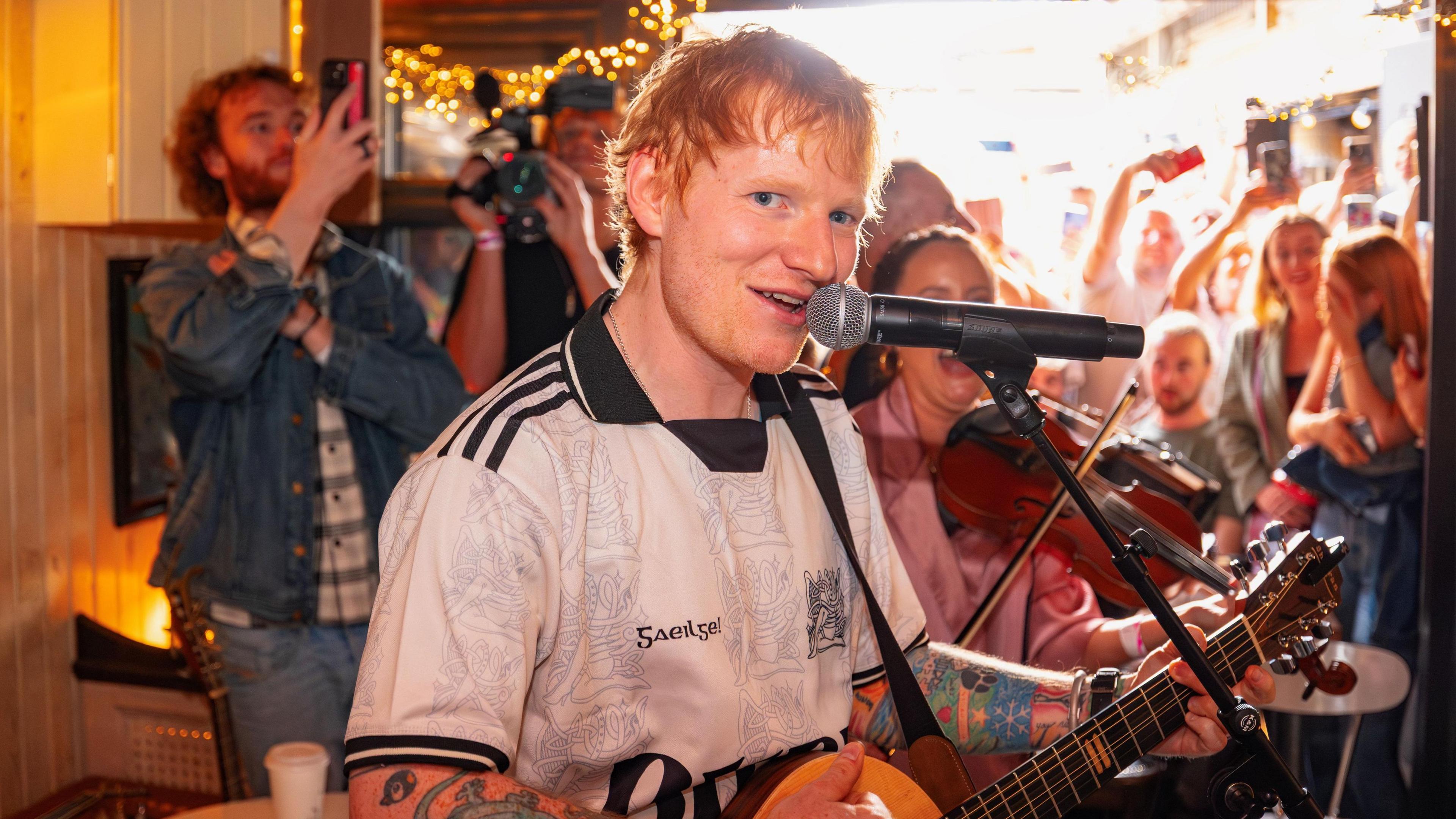Sheeran is looking into a camera while singing into a mic. He's indoors with people in the background watching and taking pictures on their mobiles.