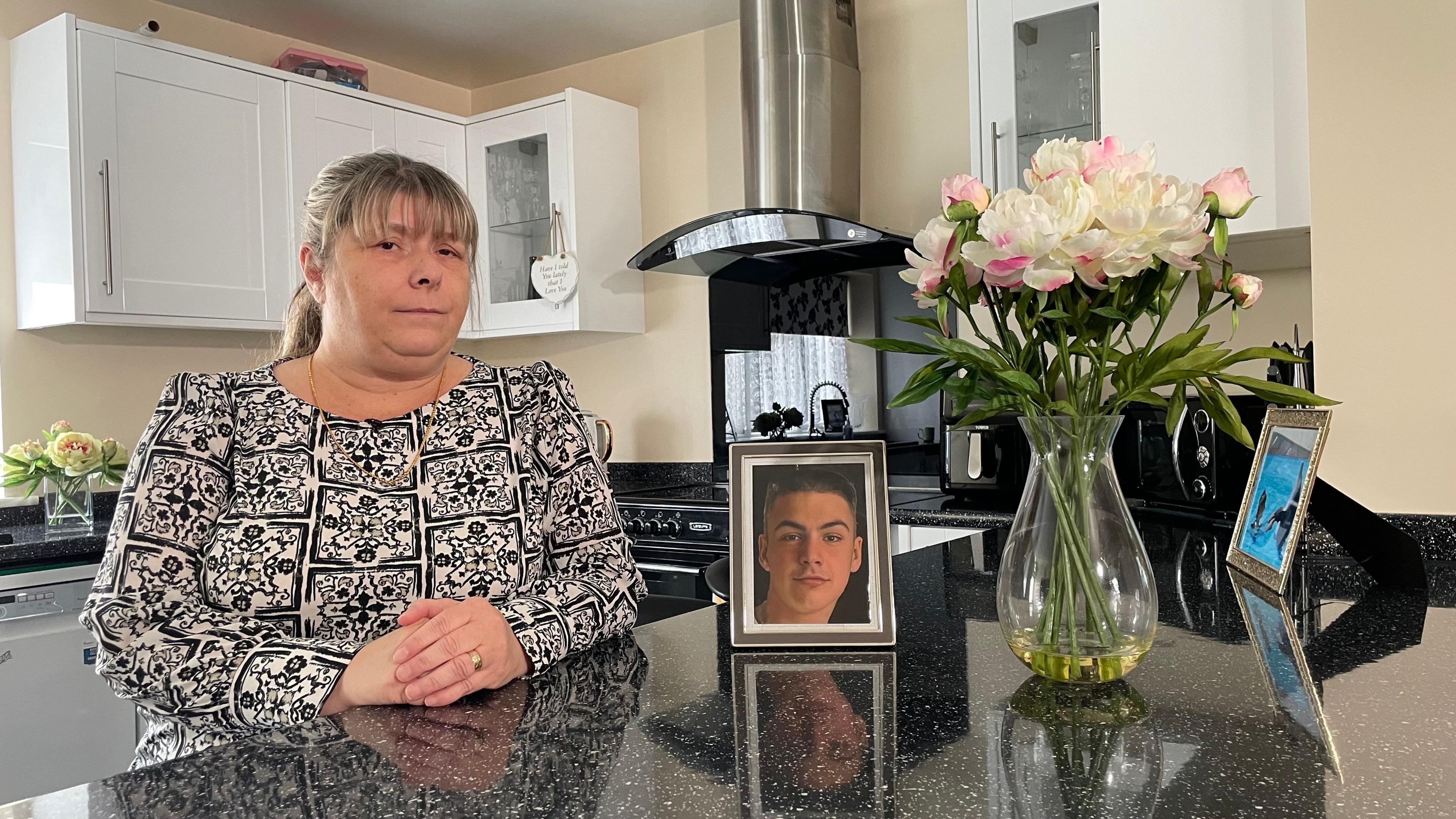 Julie Taylor is pictured in a kitchen sitting next to a worktop which has a picture of her grandson Liam on it. She is wearing a black and white patterned top and has her hands placed in each other. She has blonde hair with a fringe. A vase of flowers sits next to the photo of her grandson.