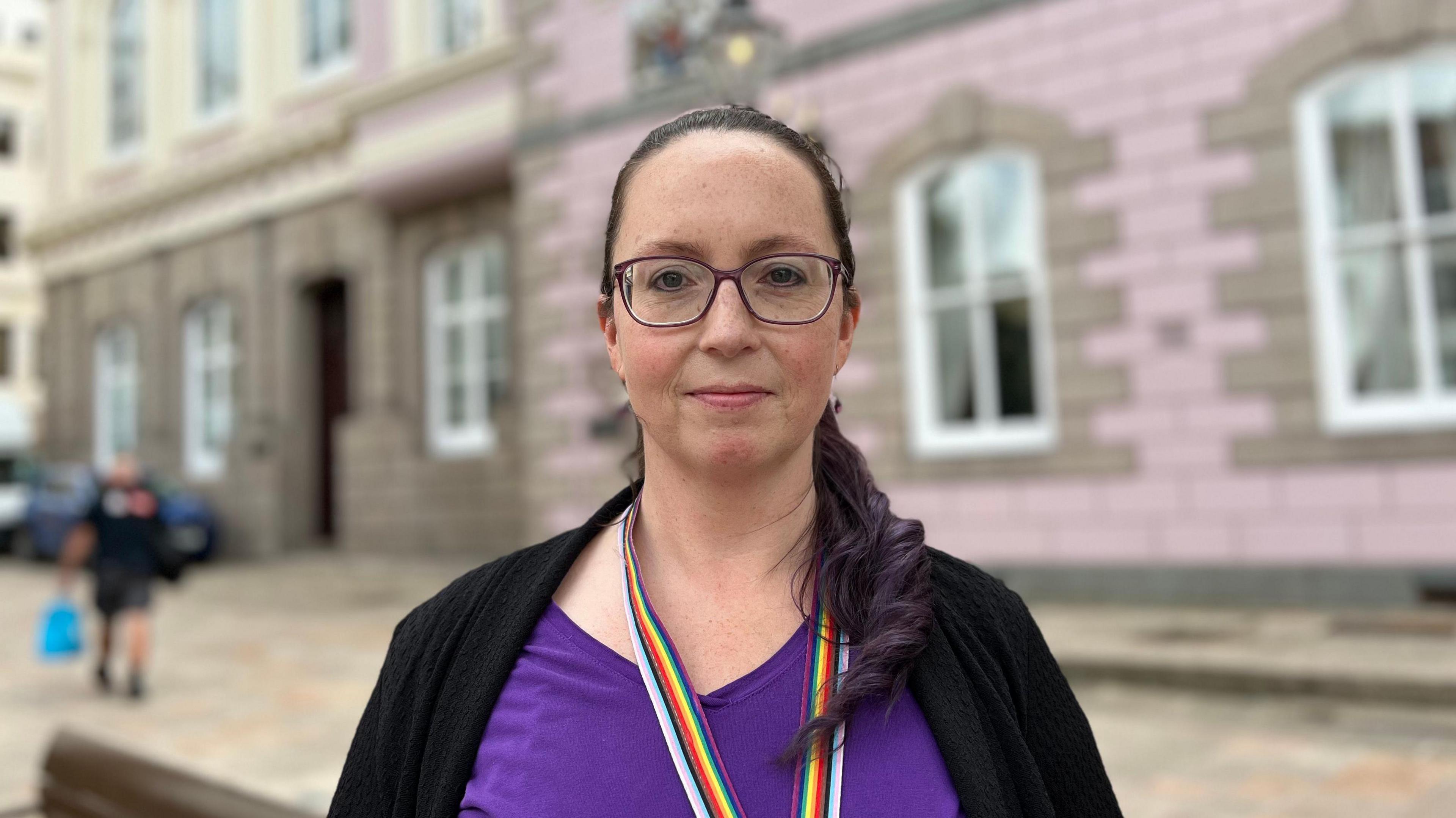 A woman wearing purple glasses, with dark eyes, looking into the camera. Her mouth is closed and black and purple hair is scraped back into a ponytail that is curled around the right side of her neck on to her chest. 