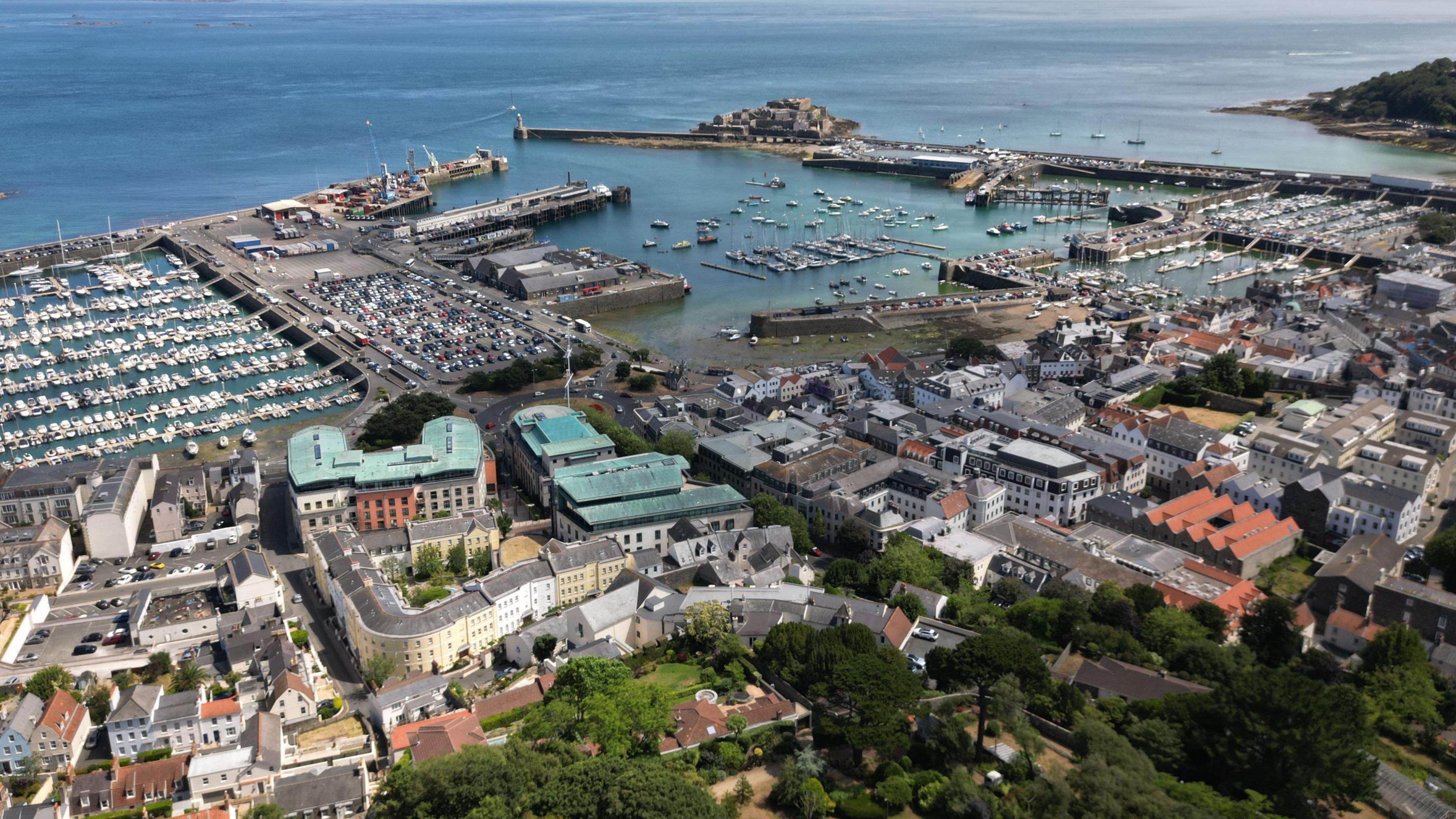 An aerial view of Guernsey on a sunny day. It shows the harbour and harbour wall in St Peter Port, with Castel Cornet on the far side.