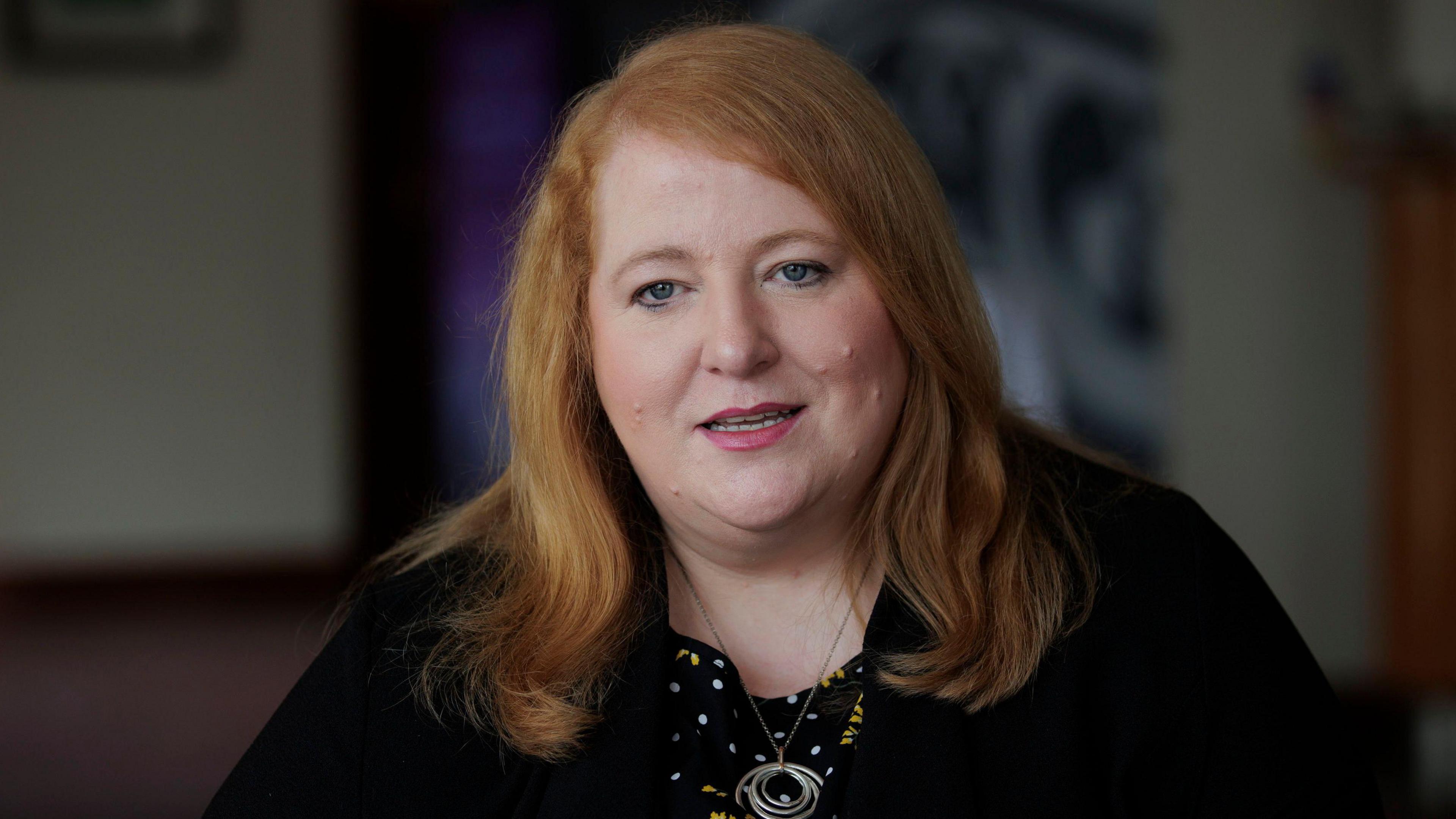 Justice Minister Naomi Long looking to camera, wearing a black jacket, a black, white and yellow top with polka dots and floral patterns, and a necklace with concentric rings. 