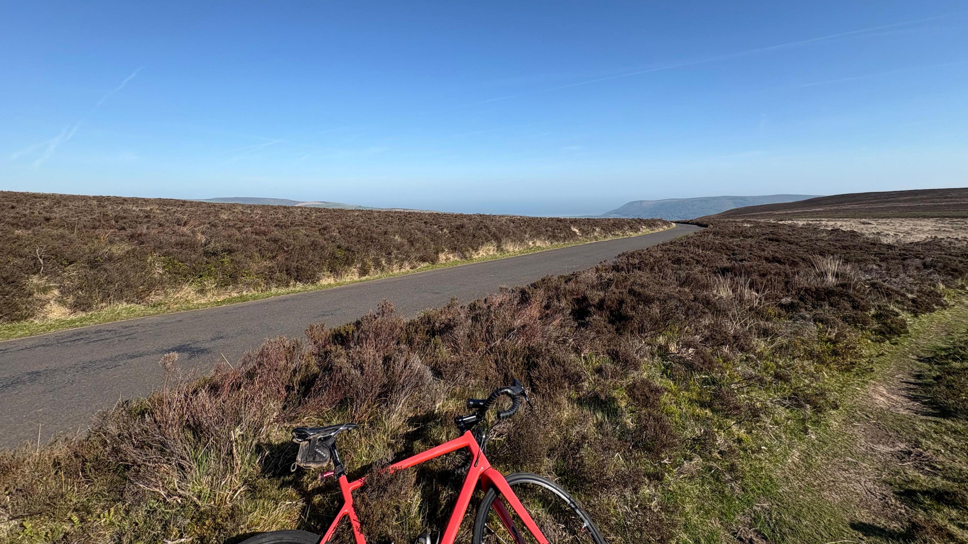 A photo of Dunkery Beacon in Exmoor, Somerset. Rolling moorland under a blue sky. A single-lane road cuts through the picture and a red bike lies in the foreground. 