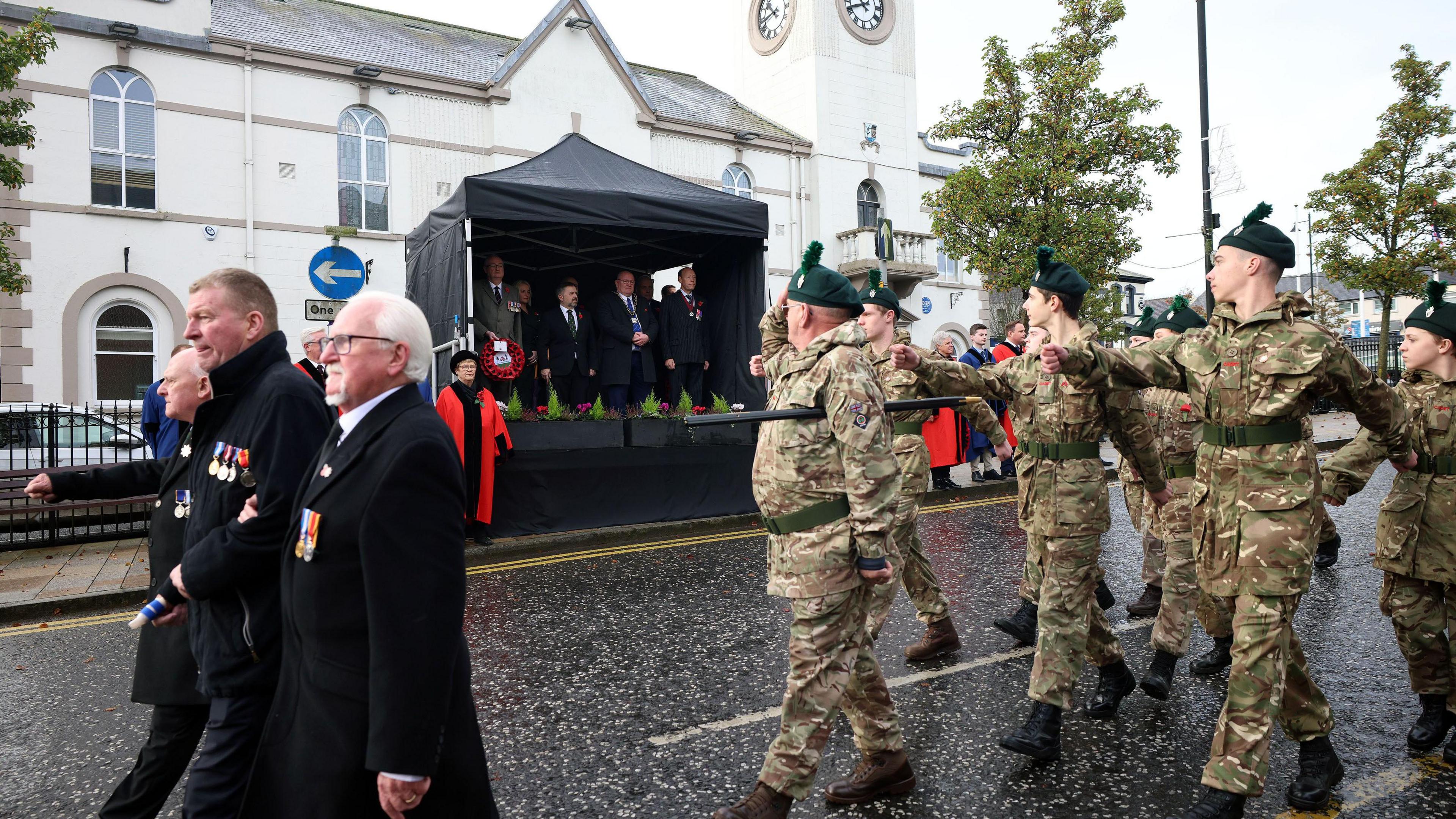 Soldiers salute as they march down a road passed a group of people standing under a gazebo.