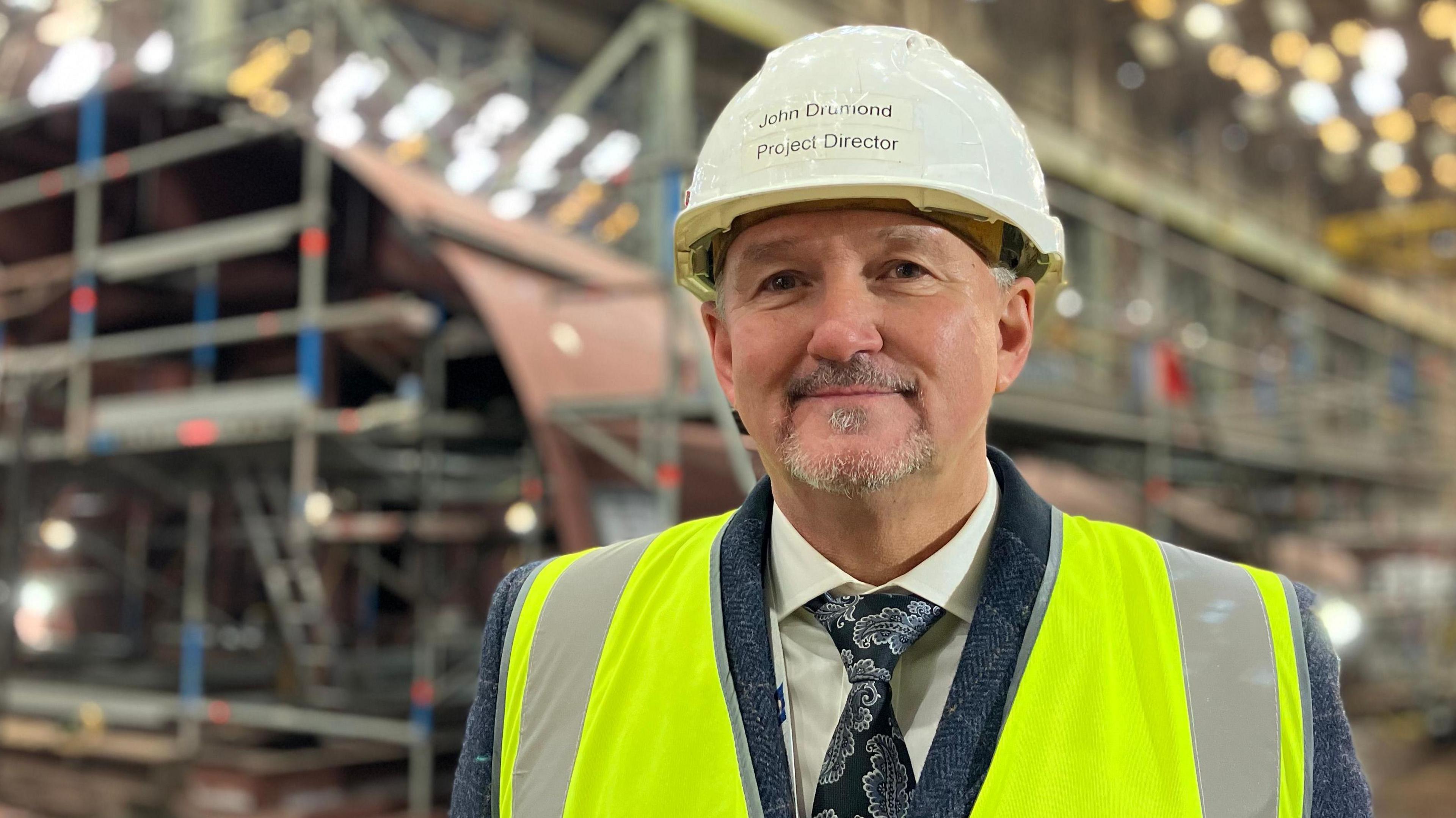 John Drummond is wearing a white hard hat and shirt an tie with a yellow high visibility vest over the top. He is stood inside Cammell Laird with the ongoing construction of the new ferry in the background.