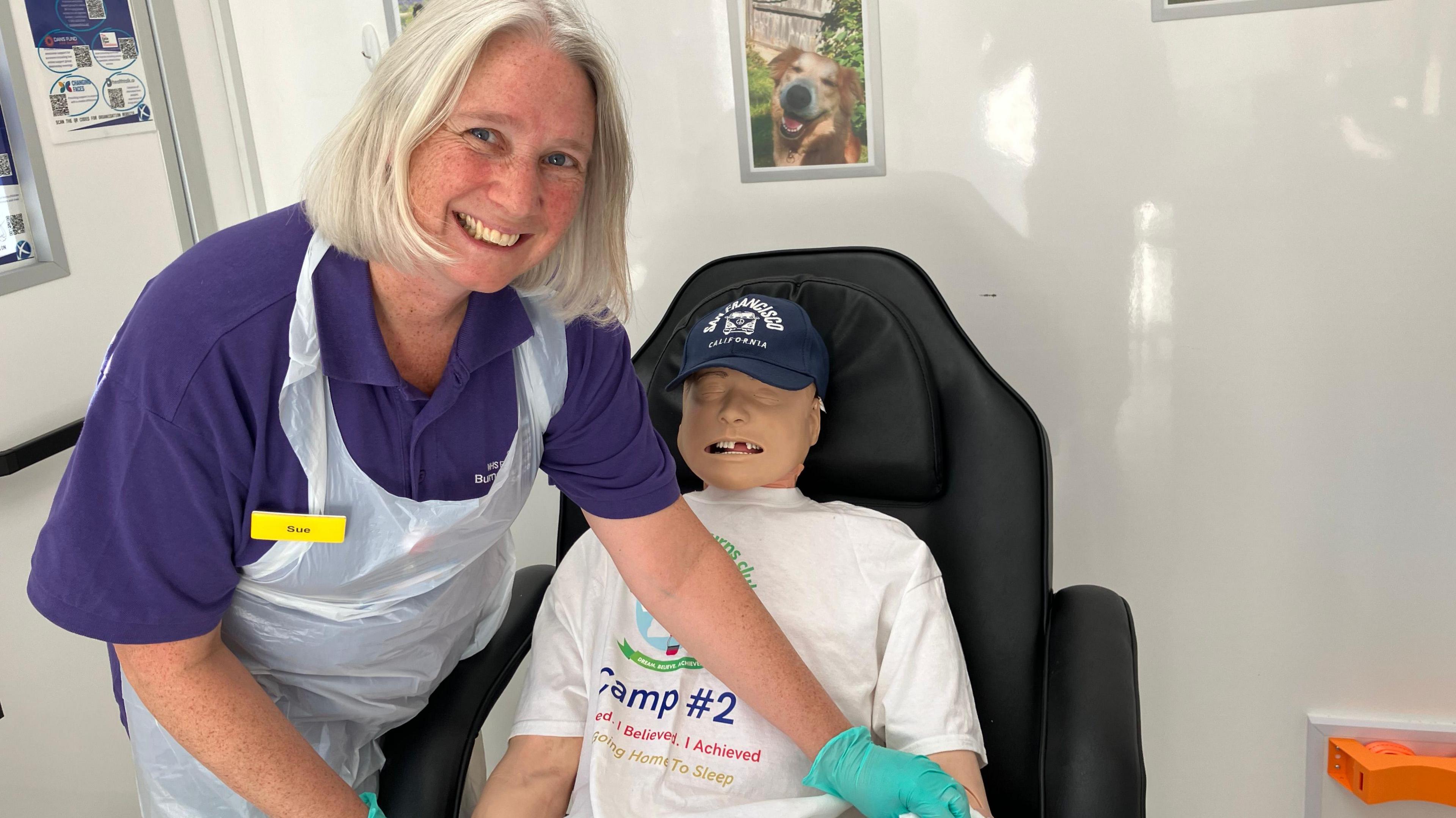A woman with short blonde hair in a purple polo shirt and a white plastic apron smiling while leaning over a dummy which is placed sitting down on a black armchair.