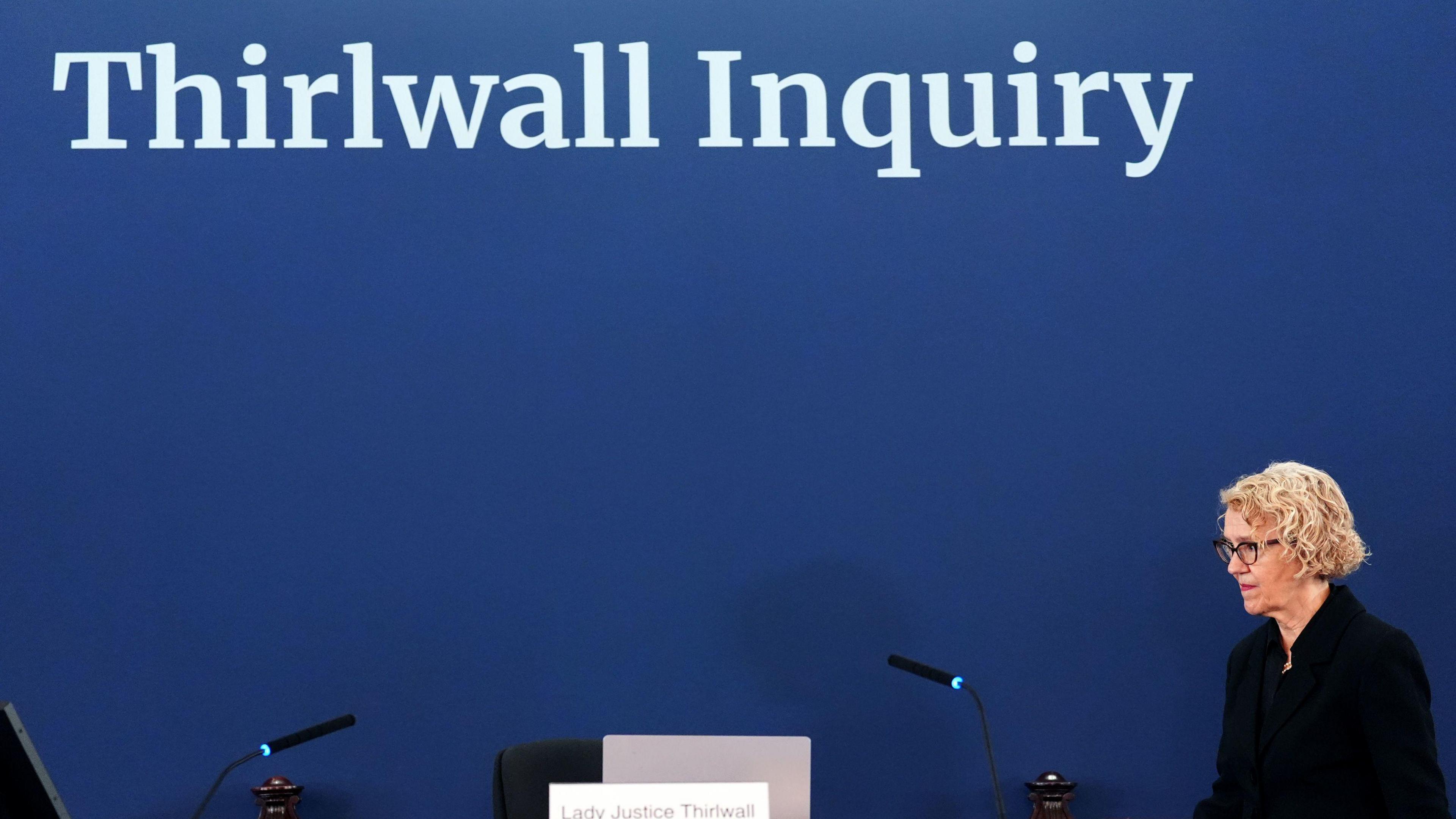 Chair of the inquiry Lady Justice Thirlwall at Liverpool Town Hall. She has wavy blonde hait and a black outfit walking on to a platform with a blue background which reads in white letters: The Thirlwall Inquiry