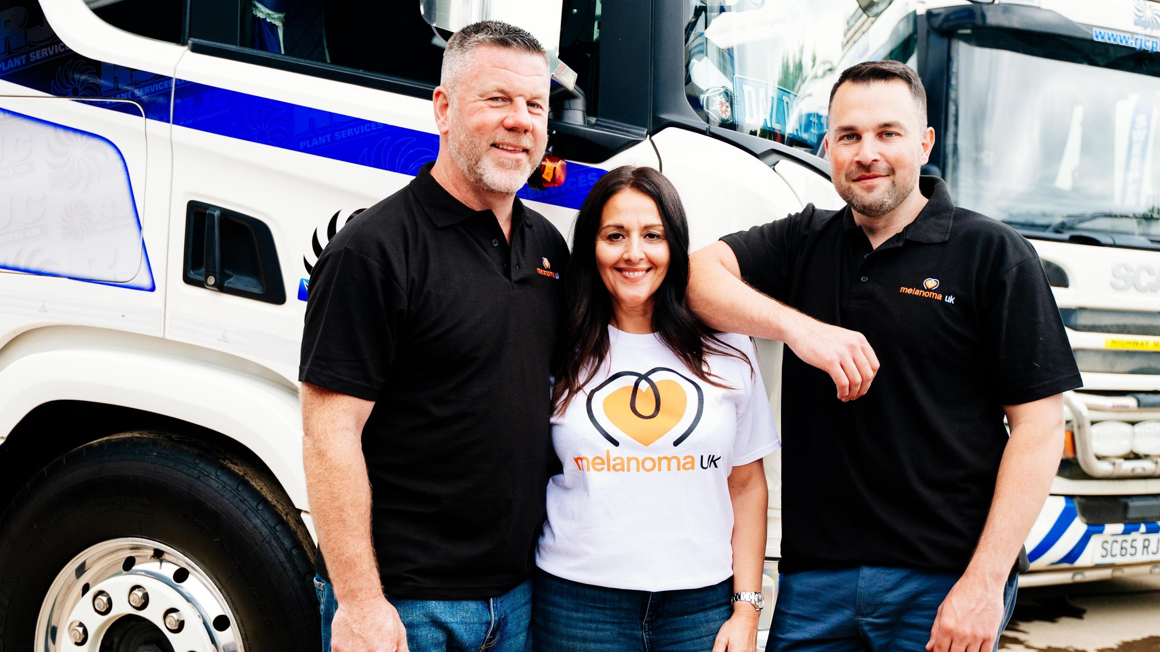 Chris Jackson, co-director, Zena Jackson, office manager, and John Harrison, co-director, stand in front of a lorry and wear Melanoma UK t-shirts