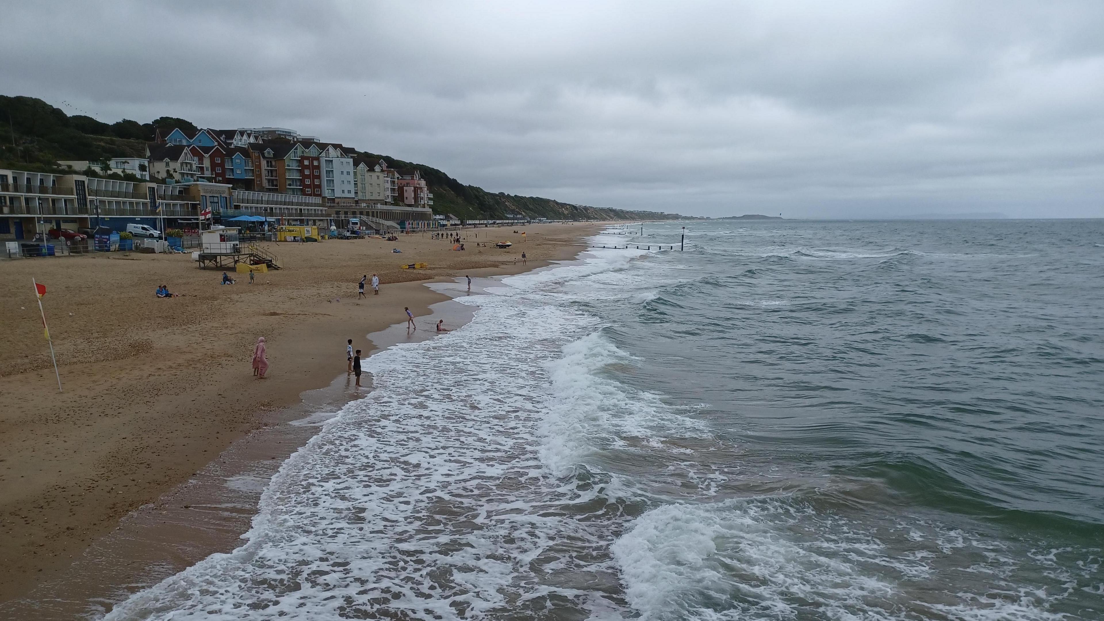 Boscombe beach on an overcast day. There are a few people on the beach with some children paddling. There are waves breaking onto the beach with white foam and the sky overhead is grey. Behind the beach there are several buildings in front of the cliff and there is a lifeguard station on the beach. 