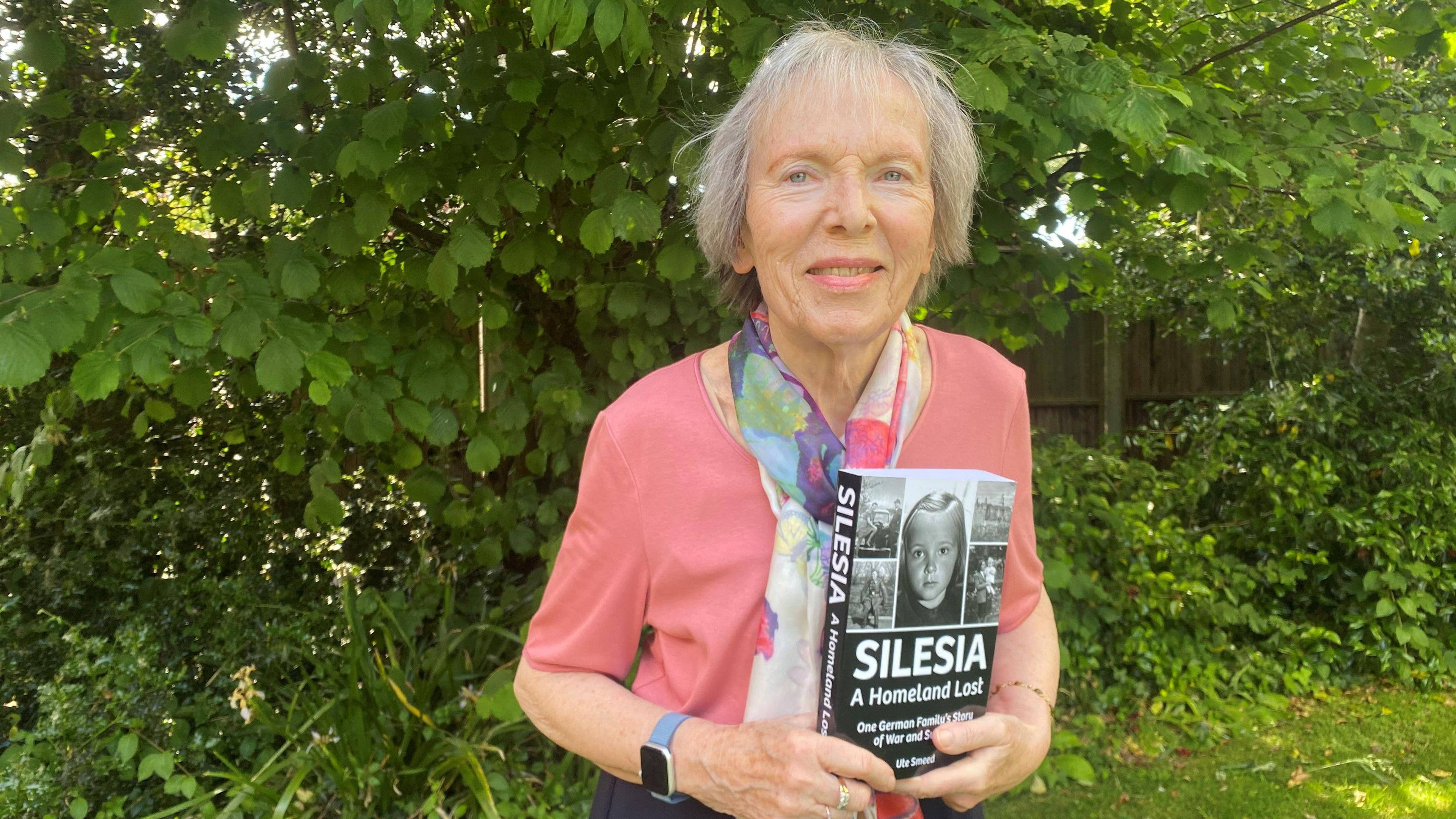 Ute Smeed, an elderly lady with grey hair. She is wearing a pink top, and has a scarf on, and is holding her book