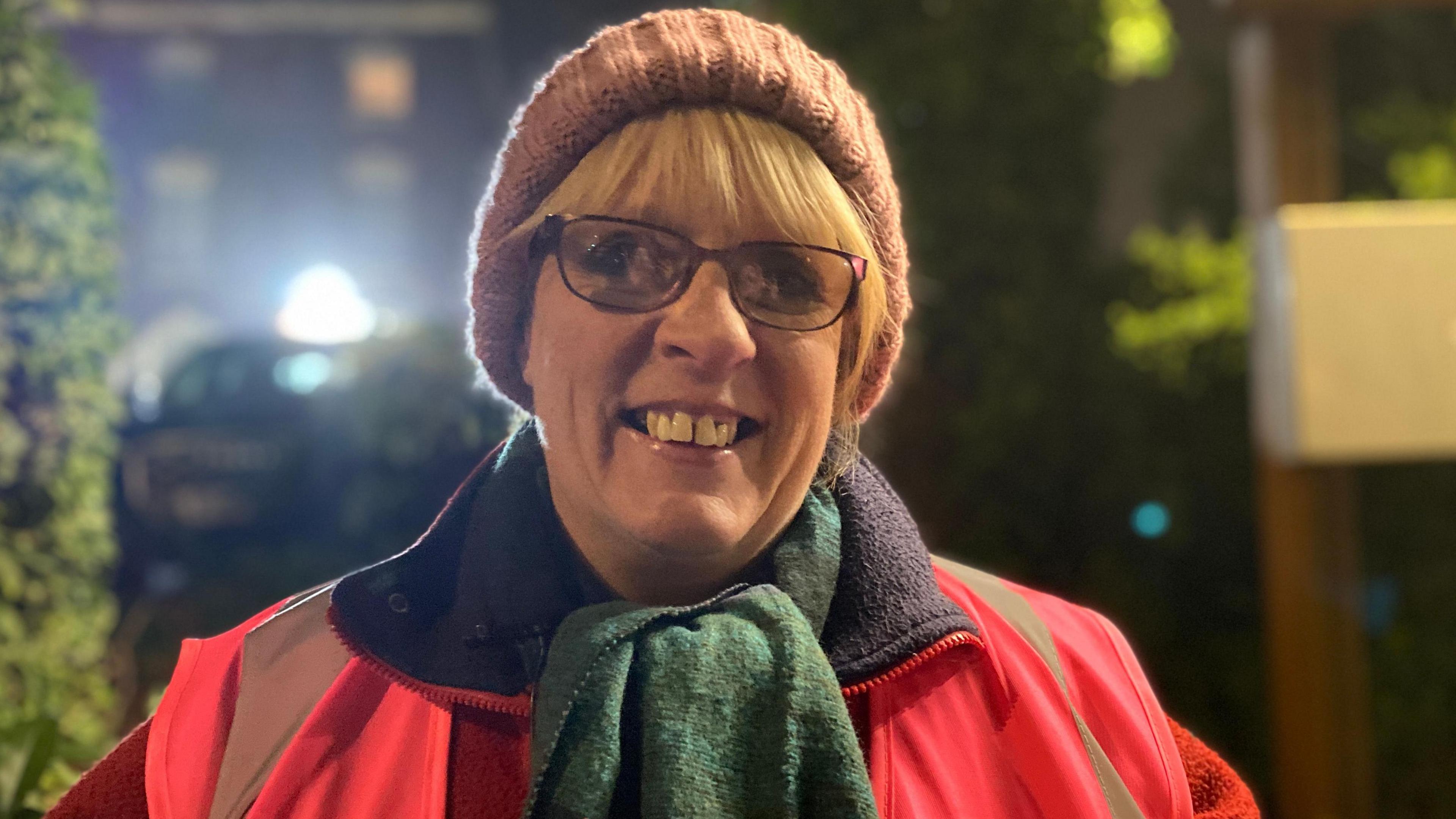 Lynda Lowe is wearing a red coat, a pink high-vis jacket, a green scarf and a pink woolly hat. She has blonde hair and dark glasses. 