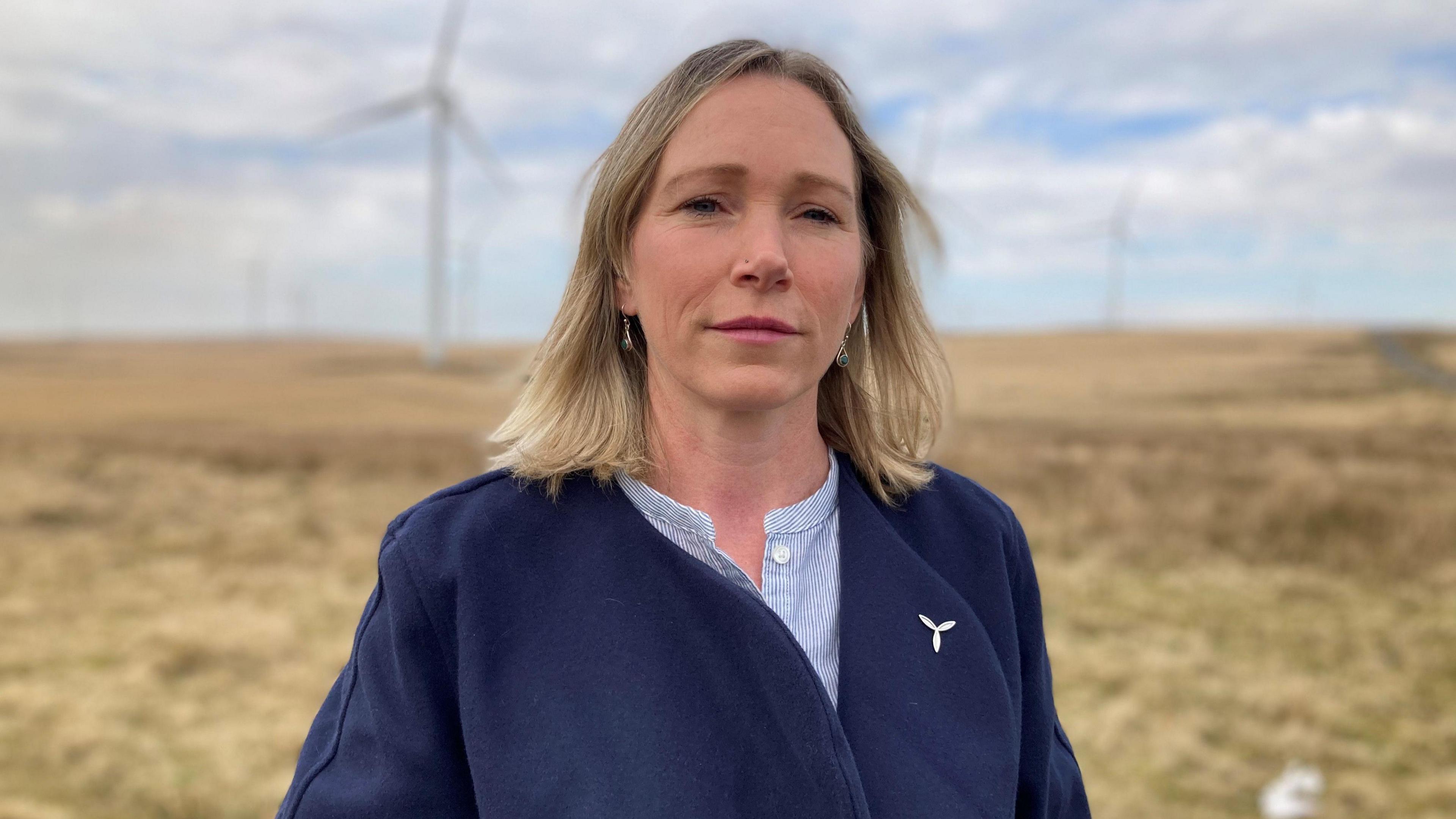 Abi Beck of RenewableUK Cymru looks straight at the camera.  She has blonde hair, and is wearing a dark blue jacket with a brooch in the shape of a wind turbine.  She is standing in a windfarm with the turbines behind her on a cloudy day.