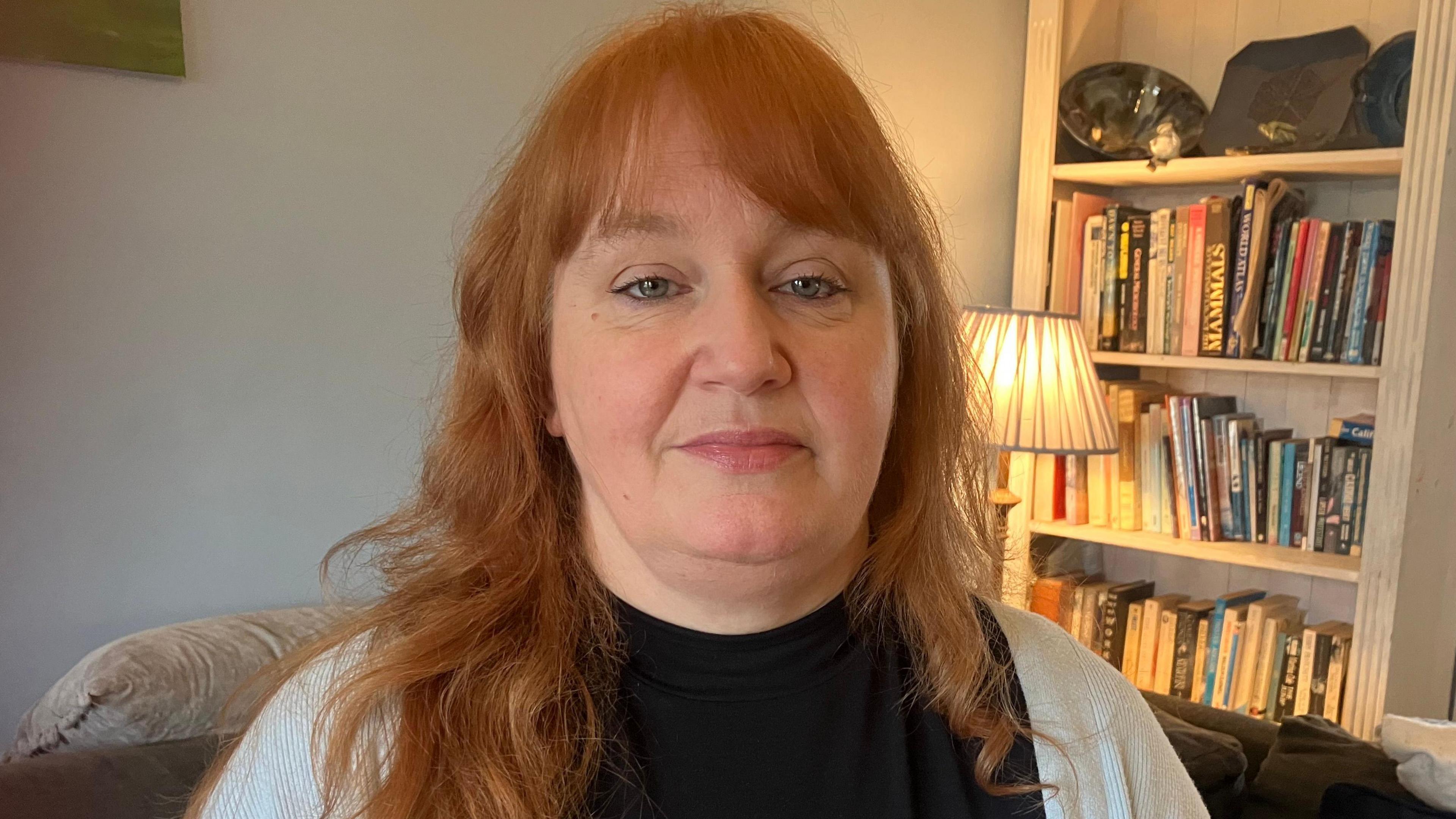 A woman with auburn hair is standing in a living room. There is a book shelf and a lamp behind her and she's wearing a black top and a grey cardigan.