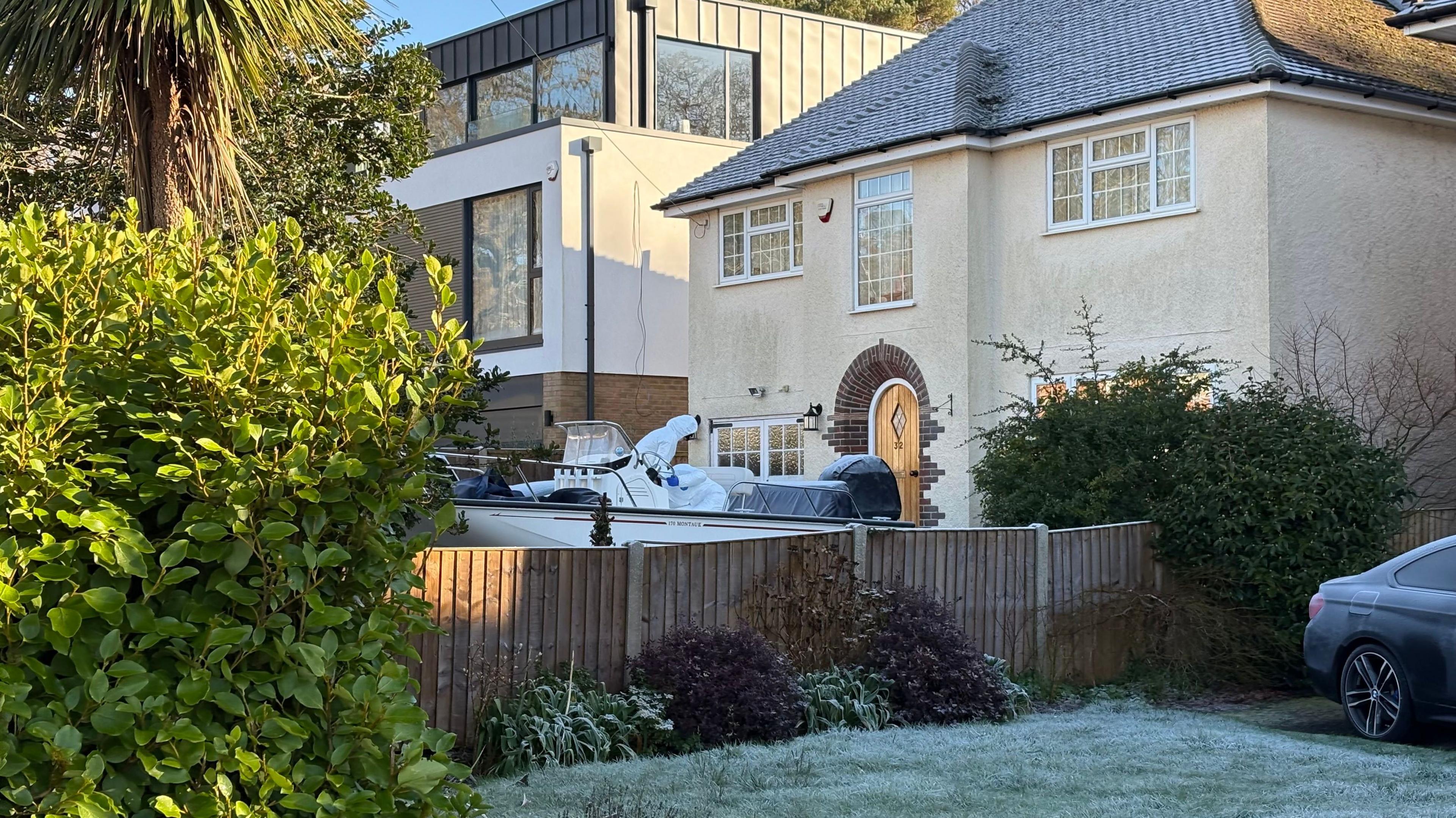 A view of a house from outside a neighbouring property shows a boat being searched on the oppiste side of a low garden fence. One person in a hooded and body length white suit can be seen on the deck of the boat. The boat is located in front of a detached house.