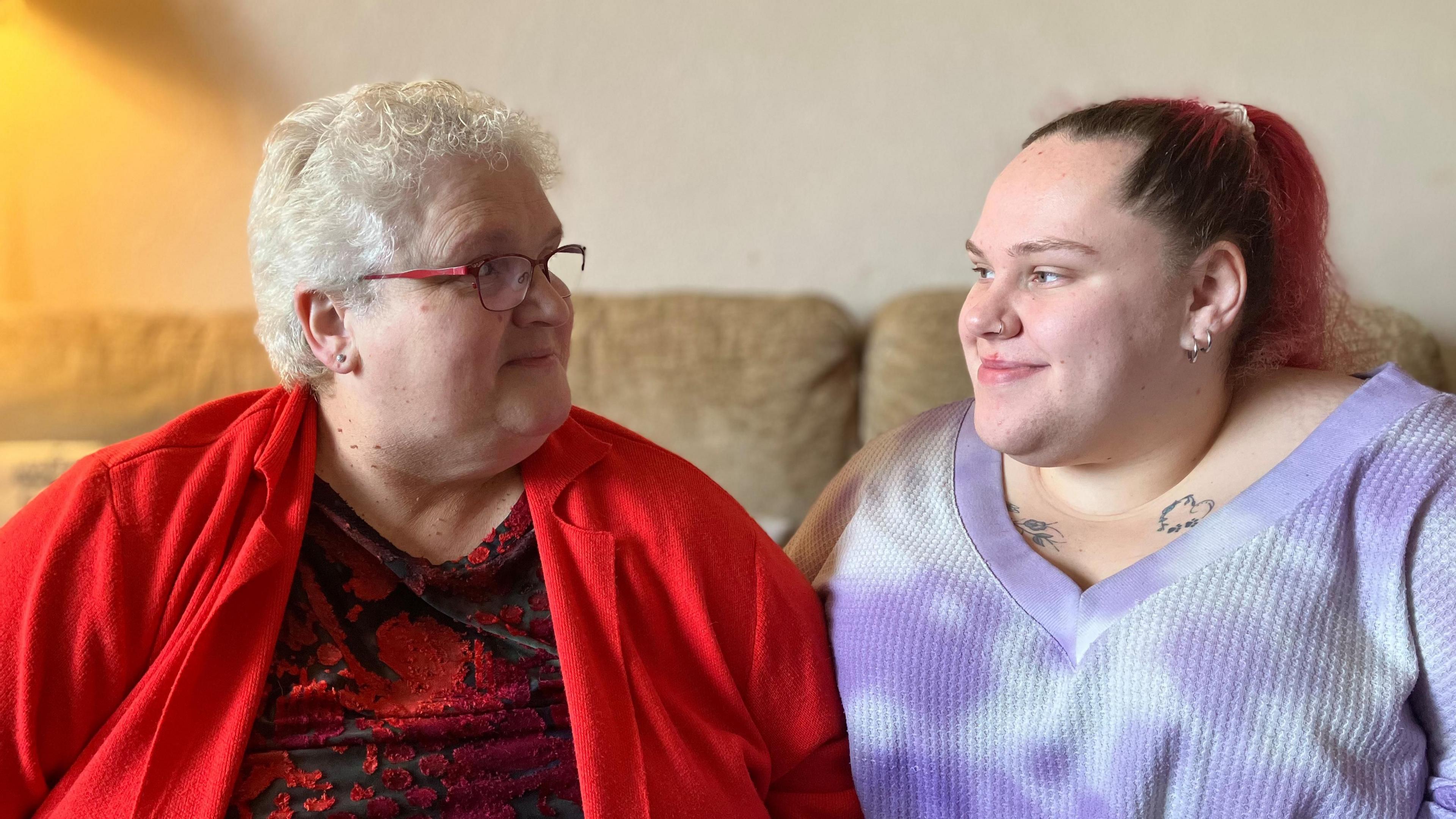 A woman in a bright red cardigan and a younger woman in a purple top sitting on a sofa together smiling at each other