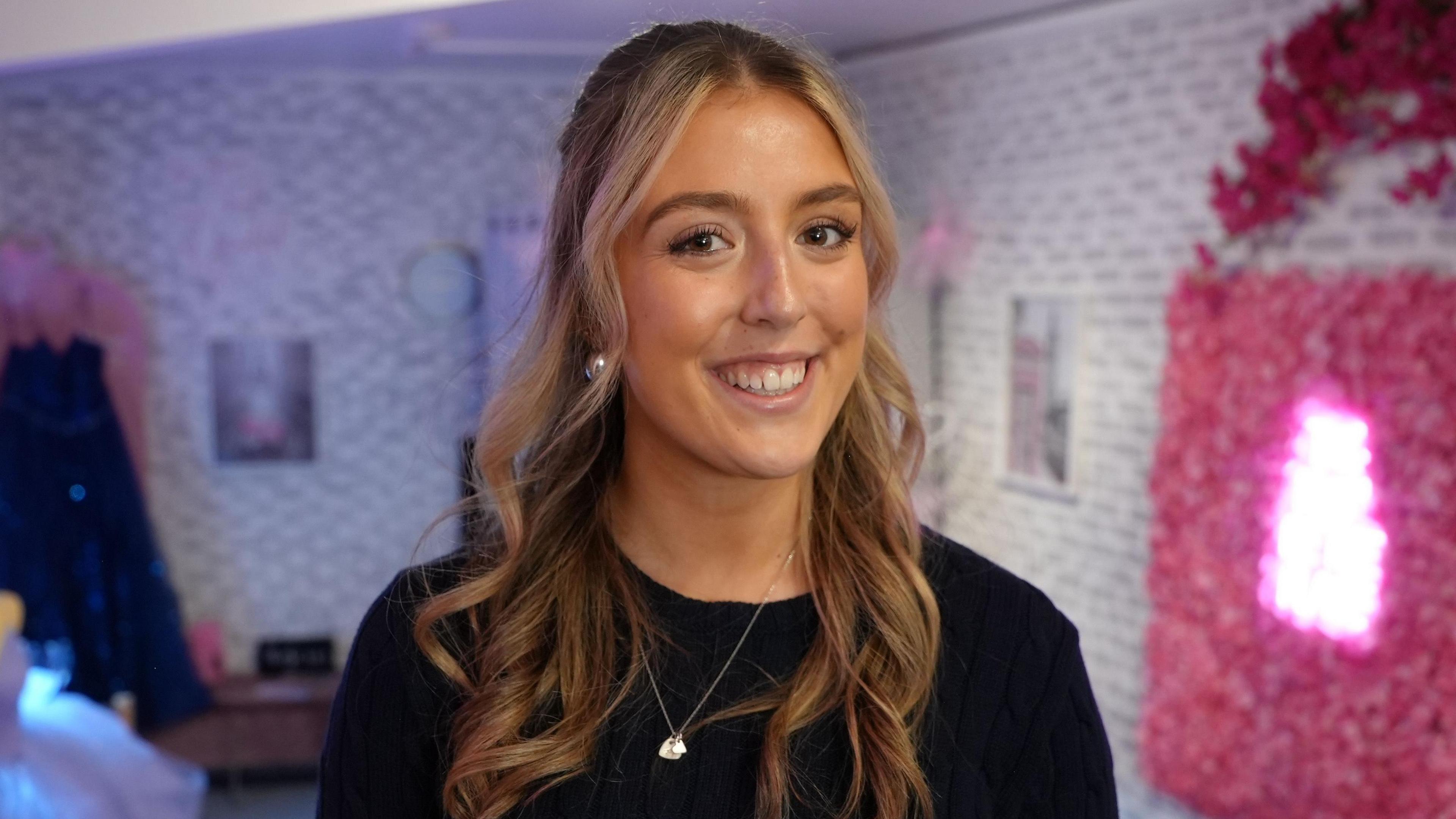 Teenage girl with long blonde wavy hair smiles, standing in the dress shop. She is wearing a navy blue round neck jumper and a silver pendant and silver earrings.  In the background is a dress hanging up on a rack and a there's a pink neon sign to her right surrounded by flower petals, but the sign is not legible in the picture. 