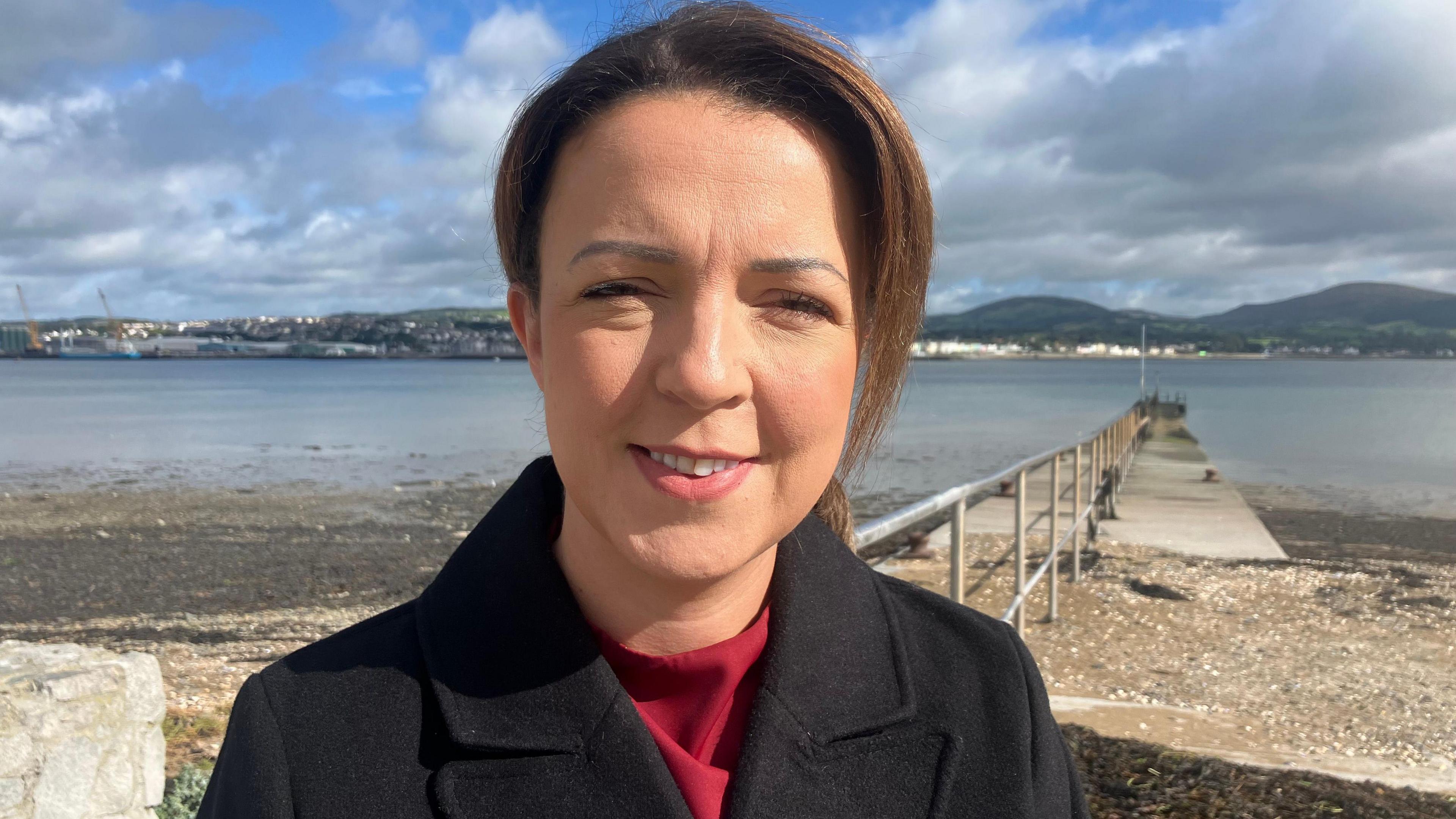 Kimmins is standing in front of a pier leading to water. She has brown hair. She's wearing a red top and a black coat.