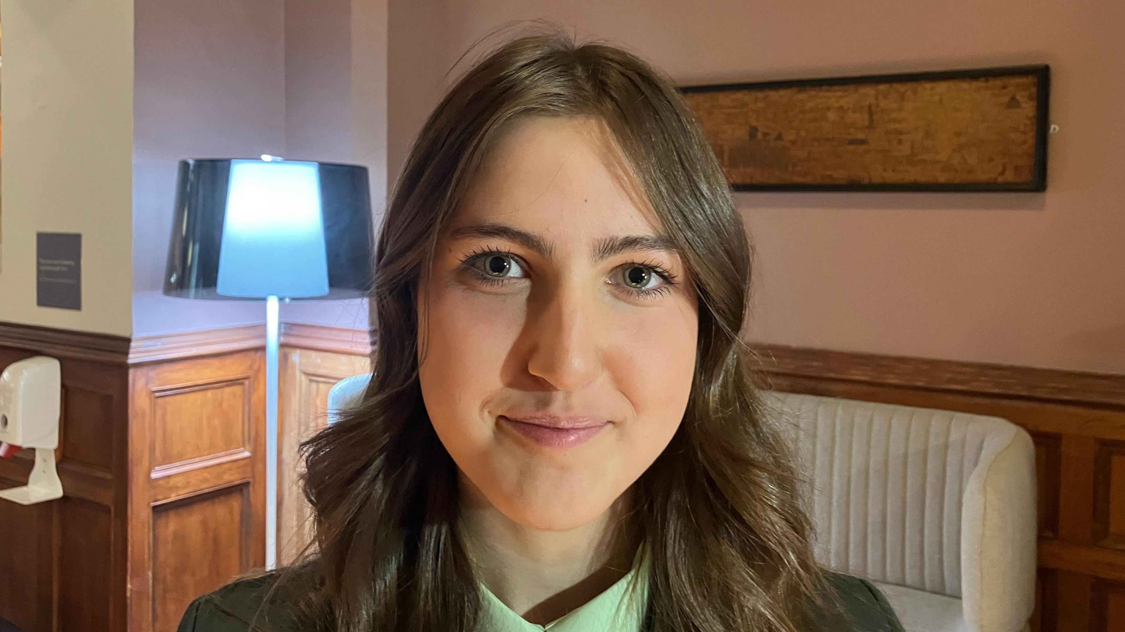 Megan has long brown hair and green eyes. She is smiling at the camera and is wearing a green uniform.