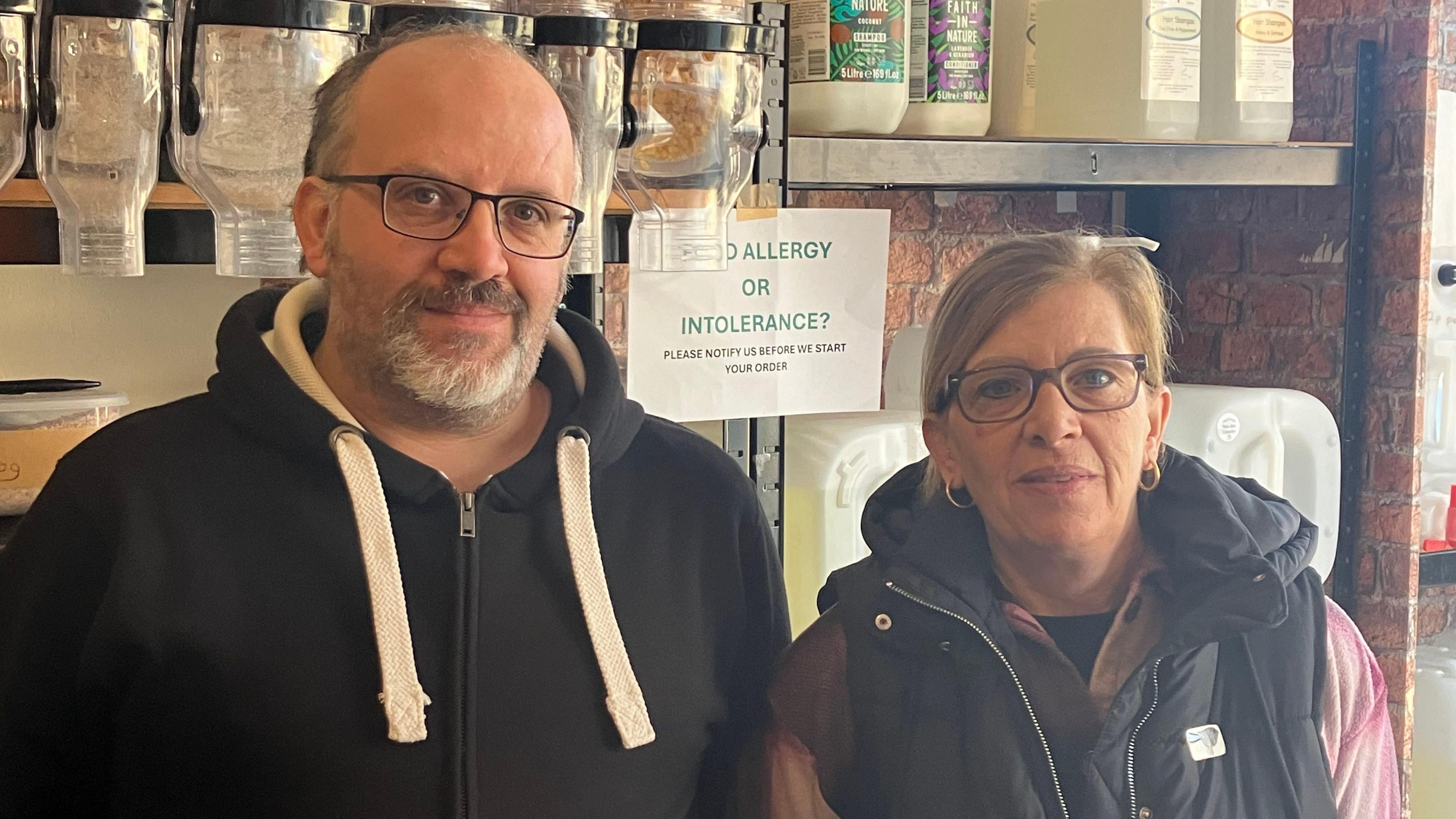 Nathan and his mother Julie Davison, both wearing black jackets and glasses, stand in front of plastic and glass containers in a shop.