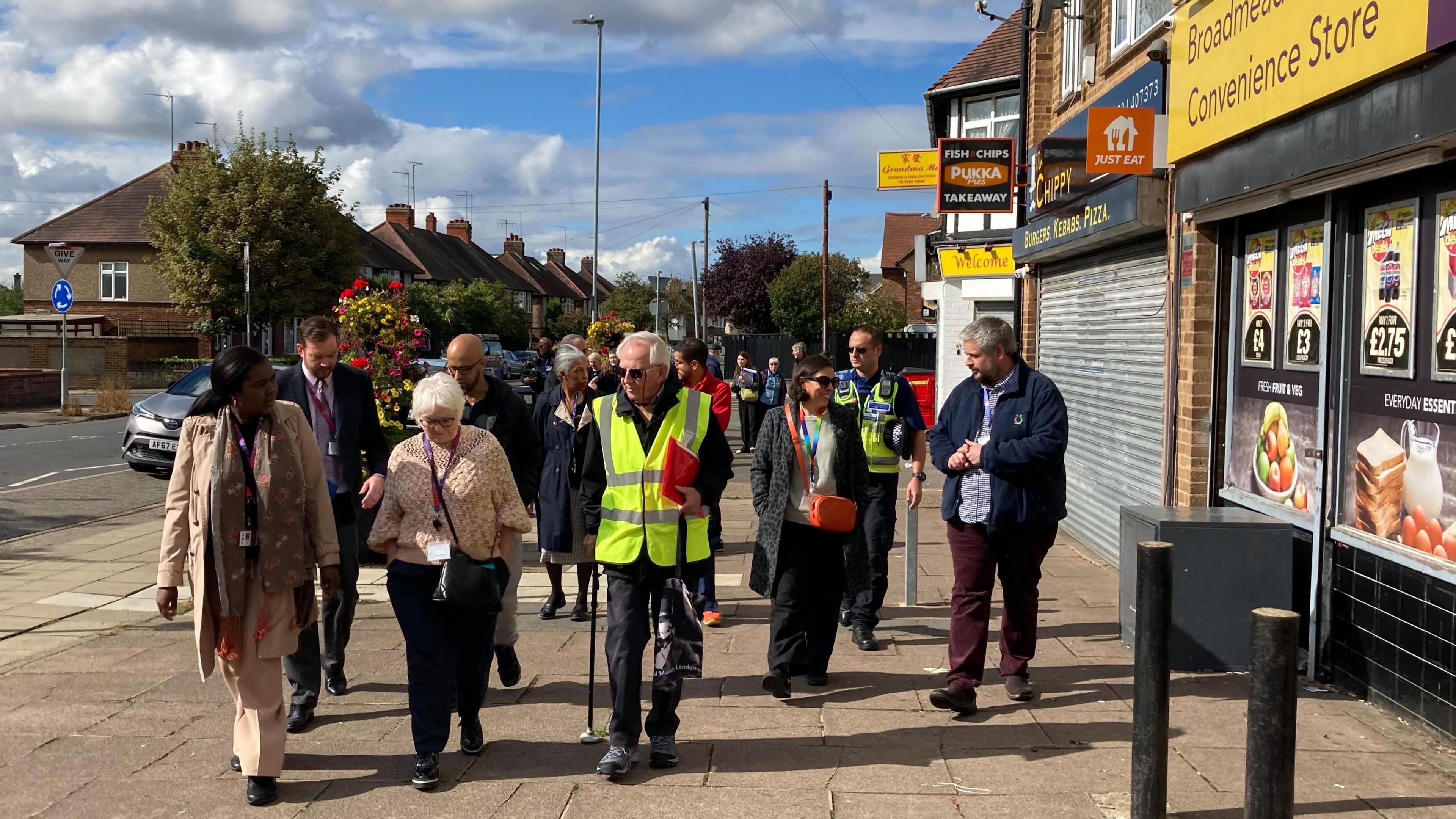 A group of people walking on a wide pavement outside a corner shop.  The front row includes a man with white hair who is carrying a red folder and wearing a yellow hi-viz jacket. Danielle Stone to his right has short white hair and a purple lanyard. A police officer is also part of the group.