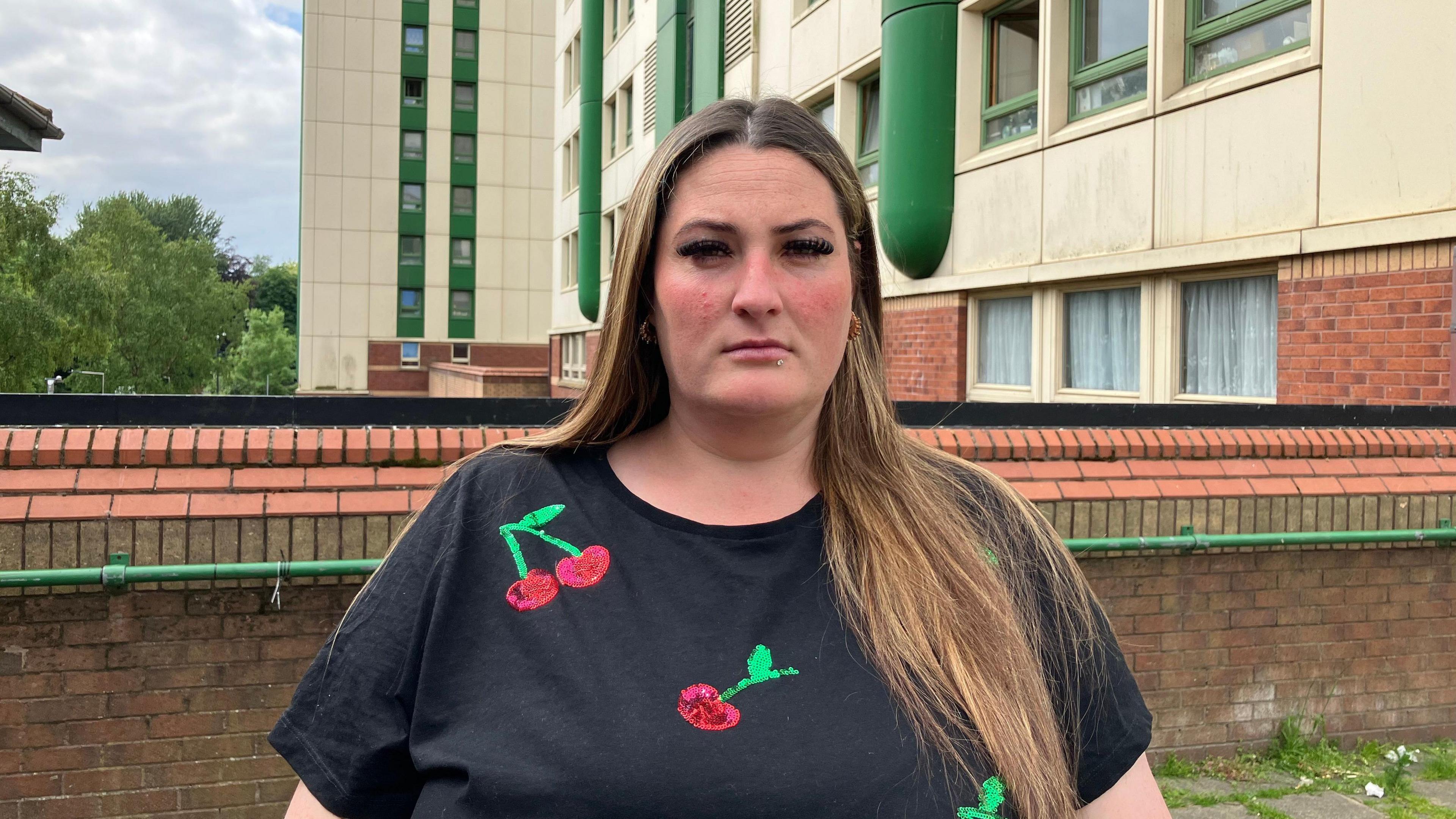 Jayne Warner is standing in front of her apartment block. She has long blonde hair and is wearing a black T-shirt with a sequinned cherry design.