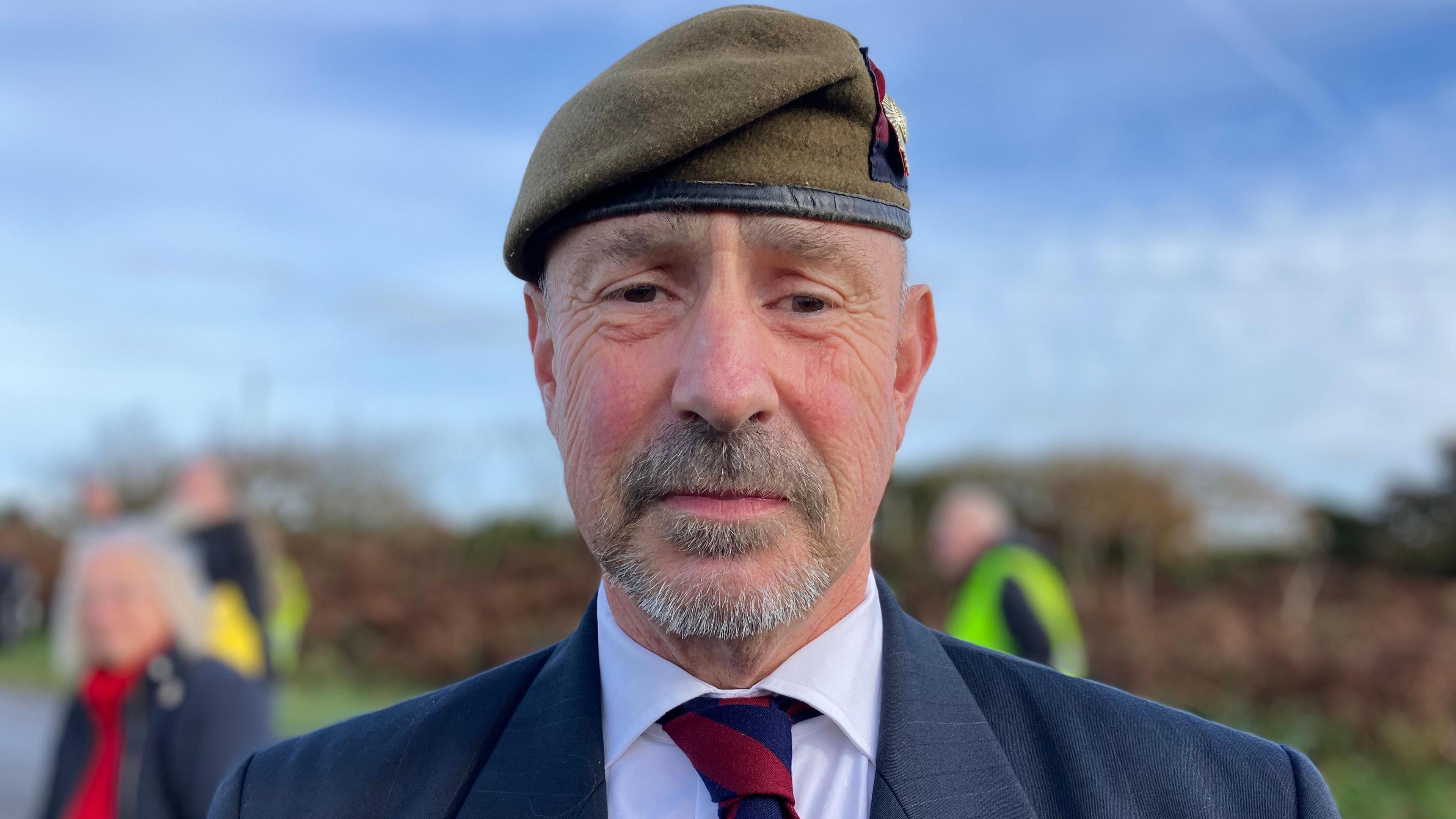 Hugh Richardson looking at the camera and wearing a dark green Army beret. He has a short beard and is wearing a white shirt with a red and two tie. He is wearing a navy blue blazer.