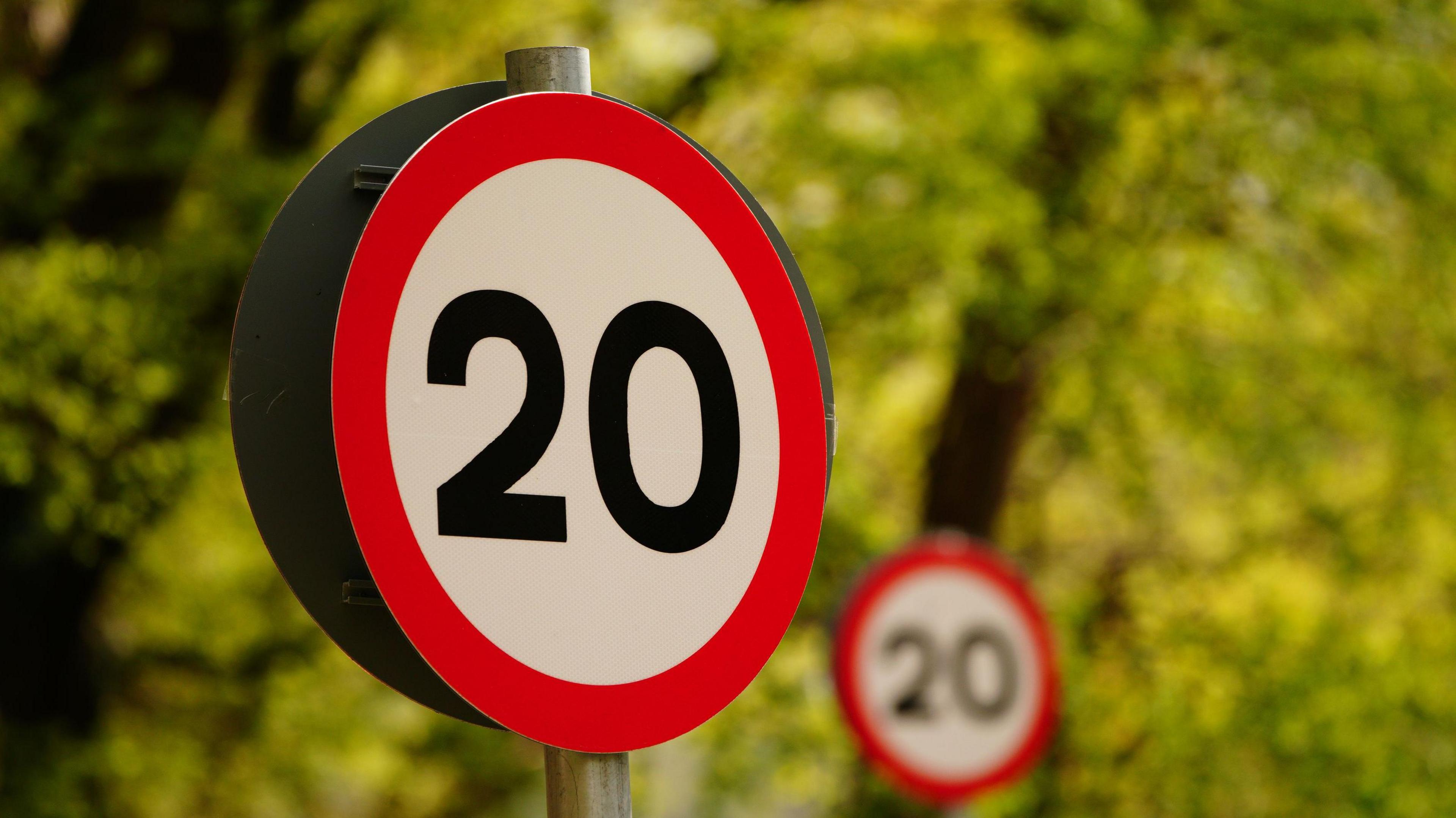 Two 20mph road signs in front of blurred trees.  