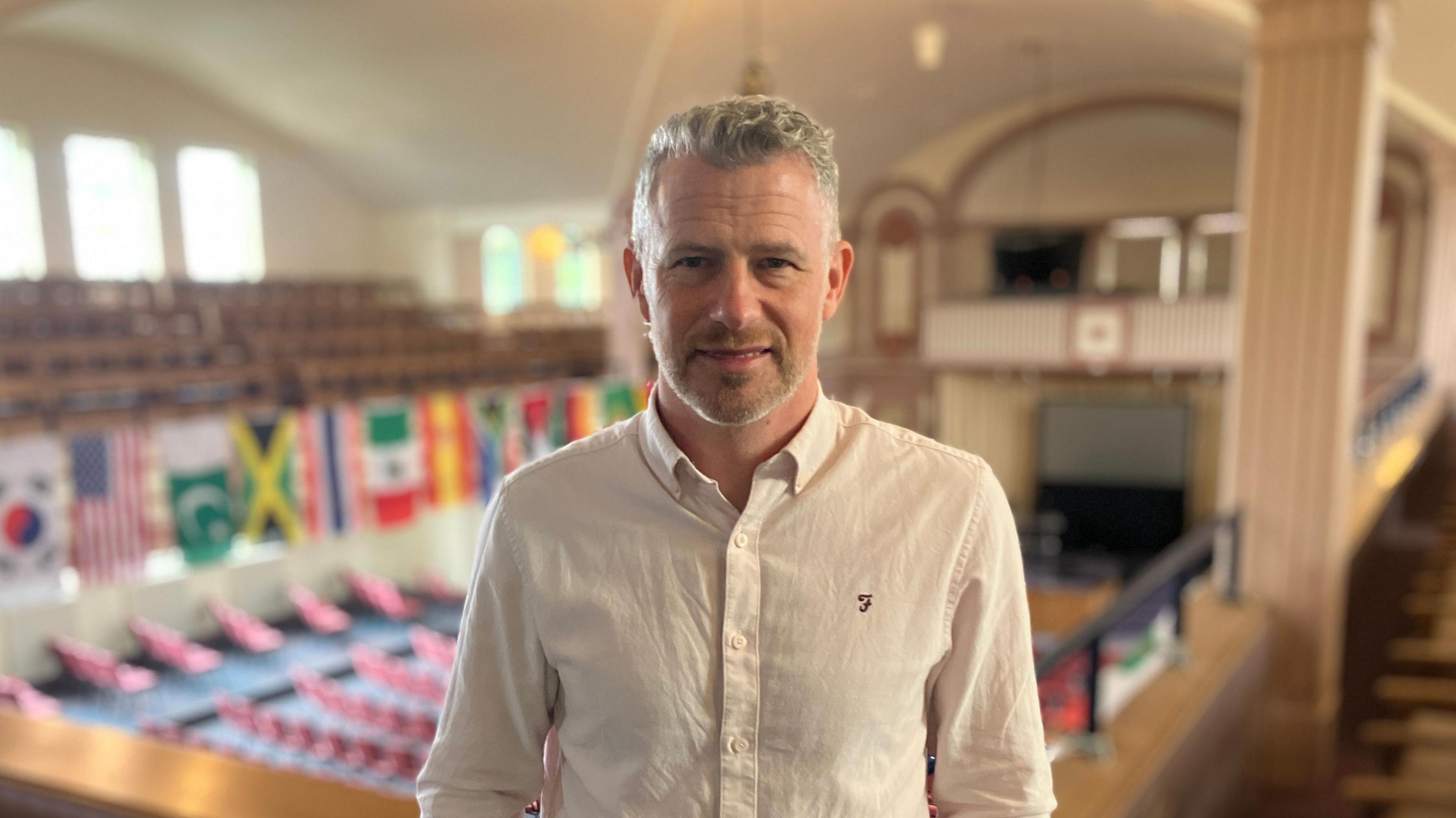 Ben Gibbins with salt and pepper hair sporting a pink shirt and a three-day stubble. In the background are several flags mounted to the wall, including Jamaica's flag.