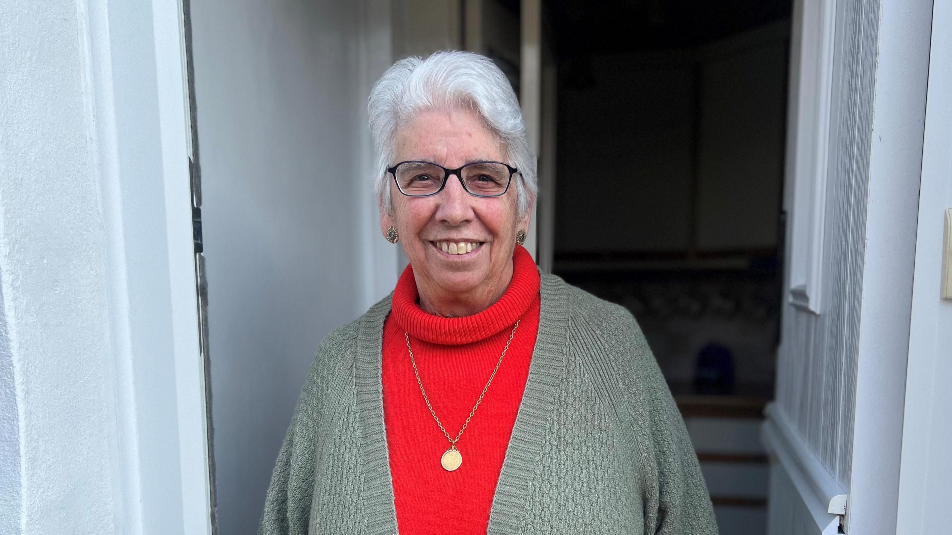 Annette Goodey has medium length white hair, and is wearing a green cardigan over a red jumper.