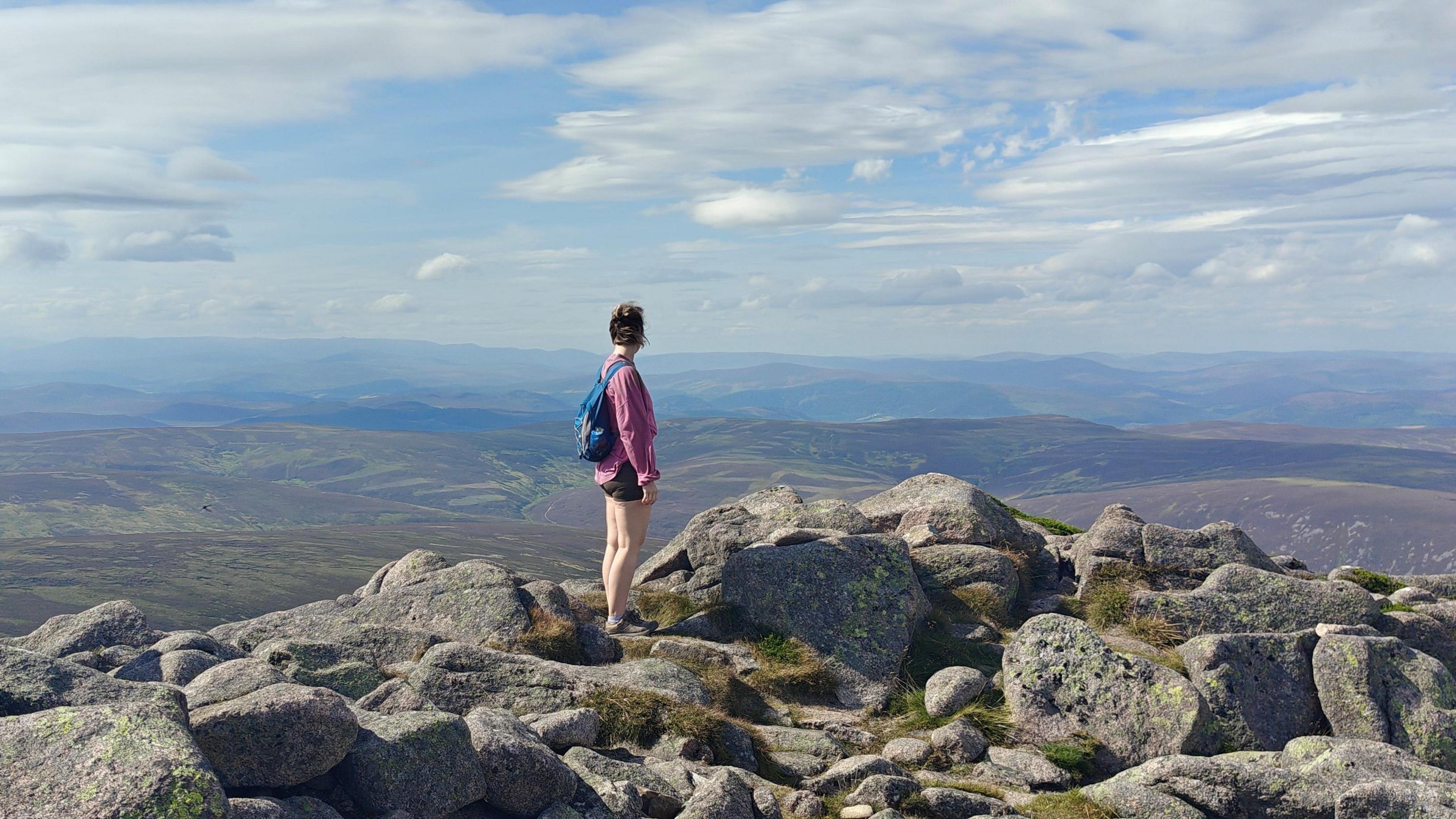 A woman wearing shorts looking into the hilly distance from a rocky hilltop.