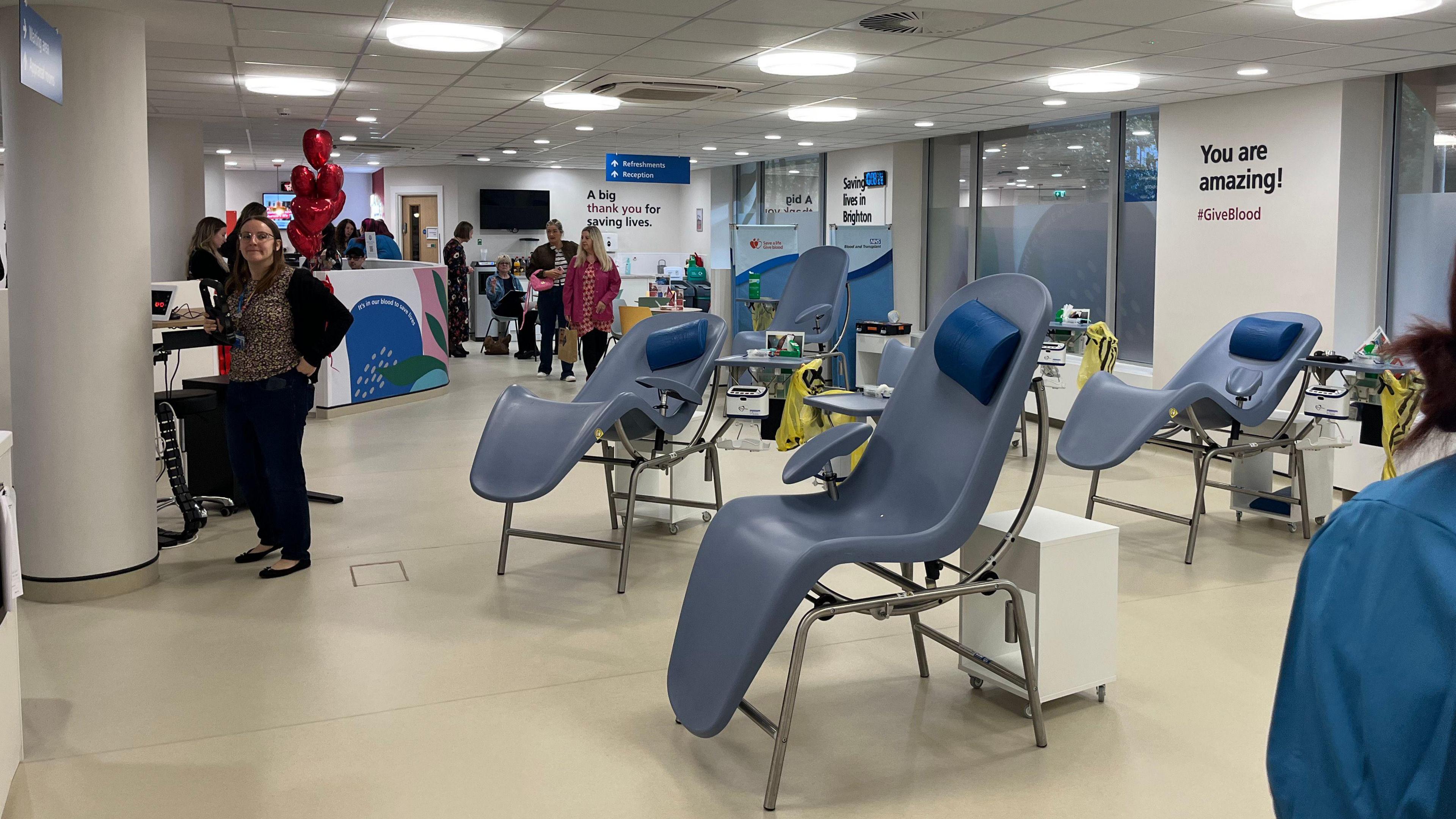 The donor centre which has lots of special chairs where people give blood. They lean back like sun loungers. People are in the background and there's balloons in the shape of hearts.