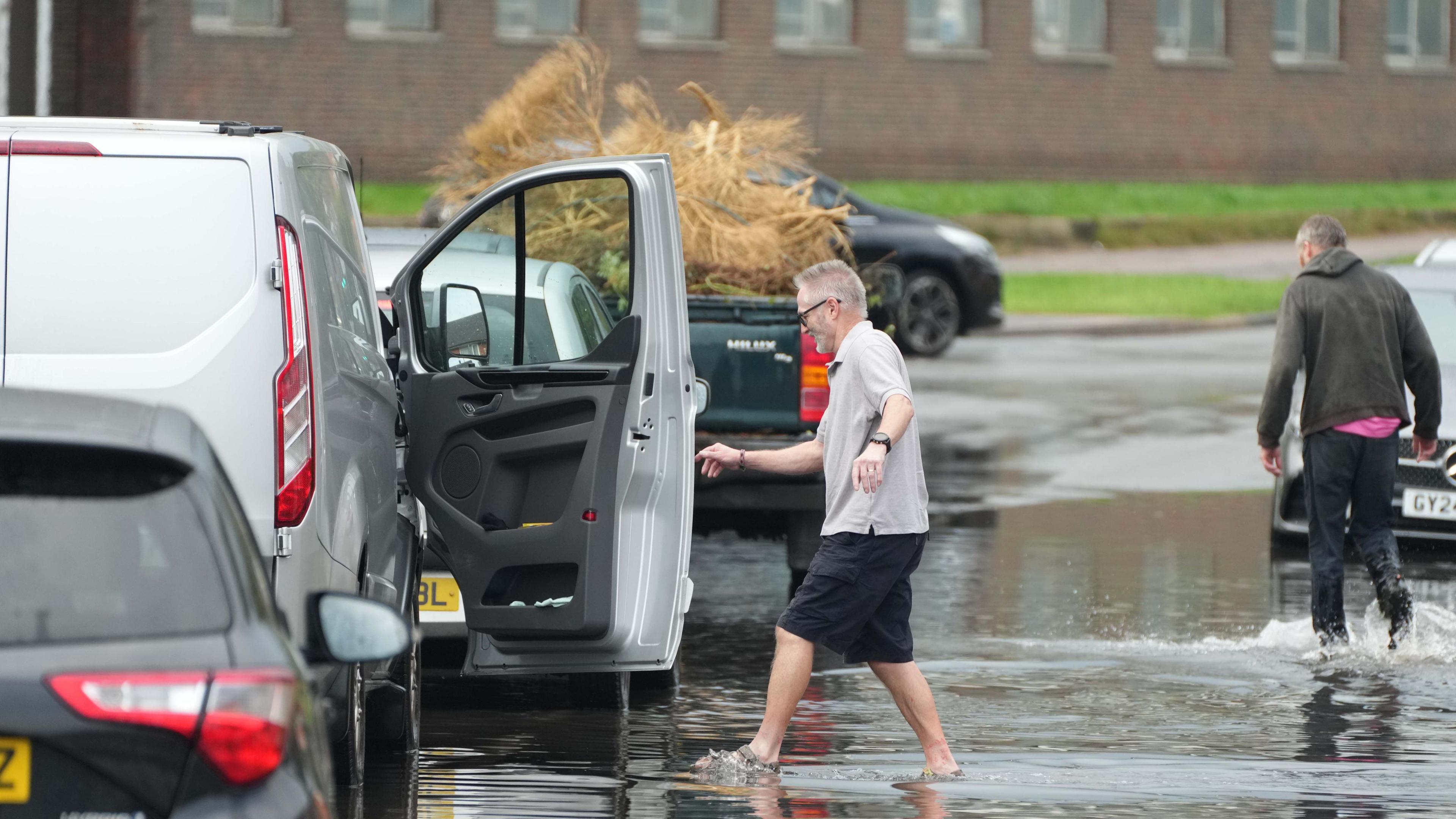 A man in shorts wades across a flooded road to an open door of a grey van.