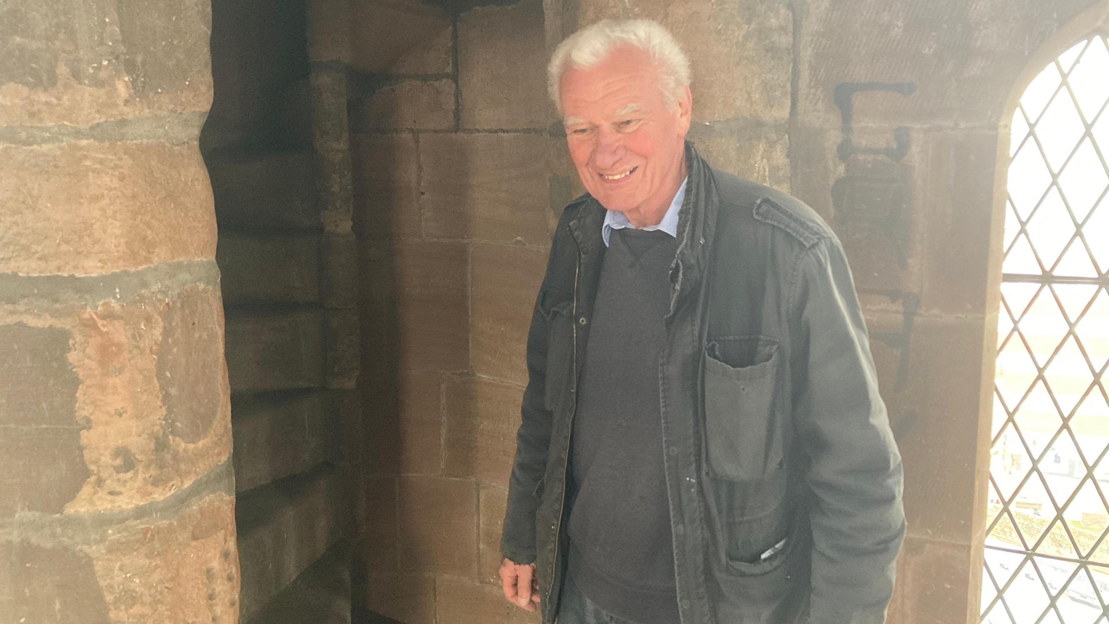 A man with white hair is smiling, wearing a grey jumper with a light blur shirt underneath and a grey jacket. He is standing inside a stone church with a glass window that has a latticed pattern on it