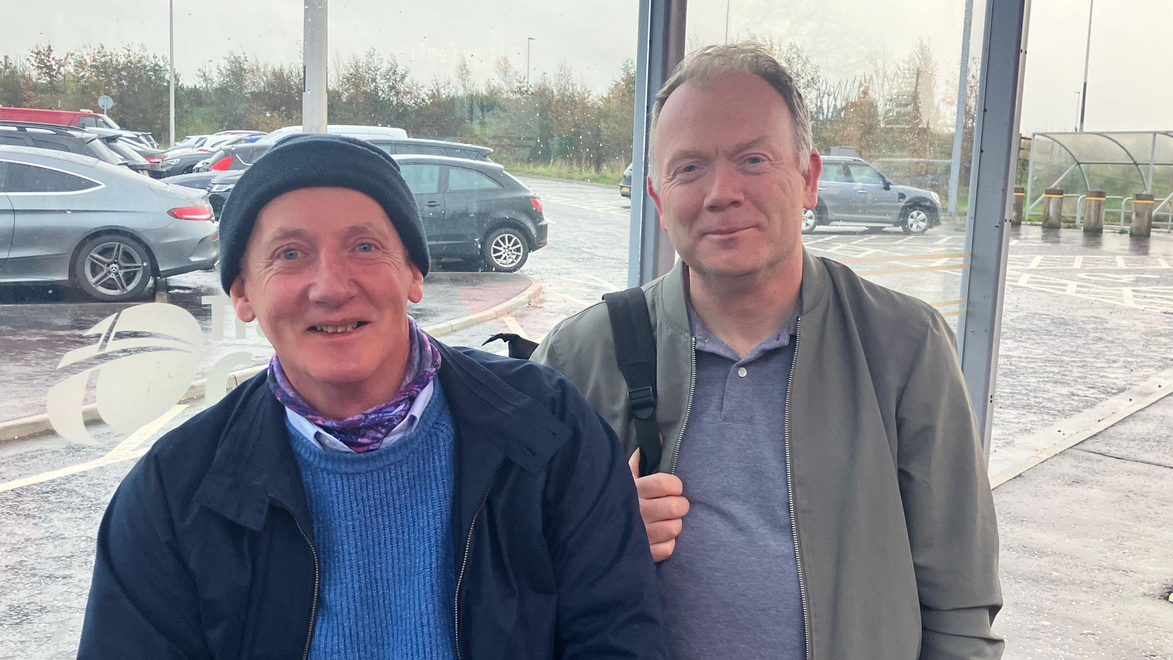 David Kelly and Kevin Harkin Are standing at a bus shelter. David has a backpack over his shoulder and is wearing a grey jacket and blue top. Kevin is wearing a blue beanie and navy jacket.