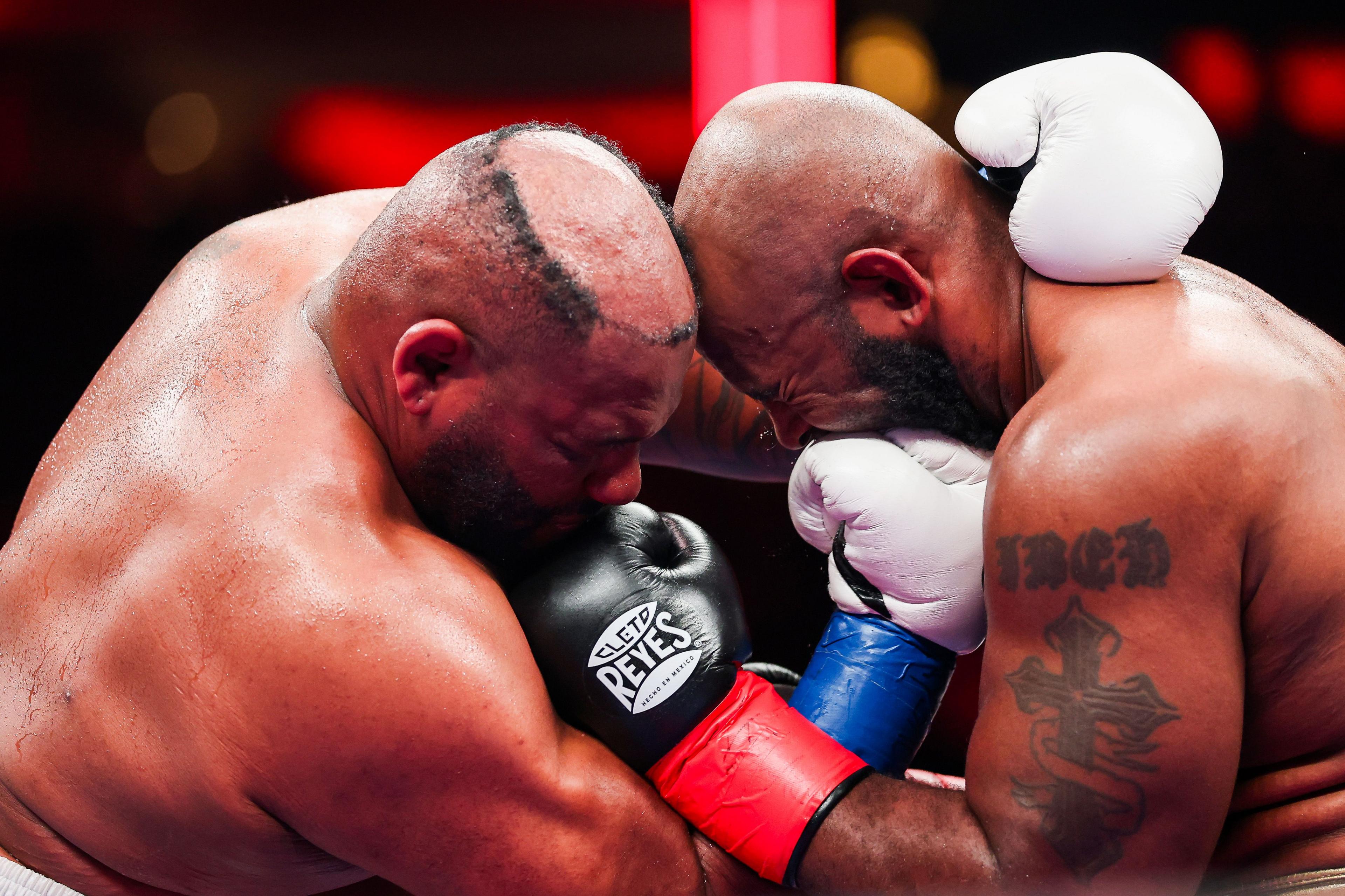 Boxers Jarrell Miller and Kingsley Ibeh lean in close during an intense exchange, their gloves pressed against each other as they battle at close range. One boxer wears black gloves and the other white, with both fighters straining with effort under bright arena lights.
