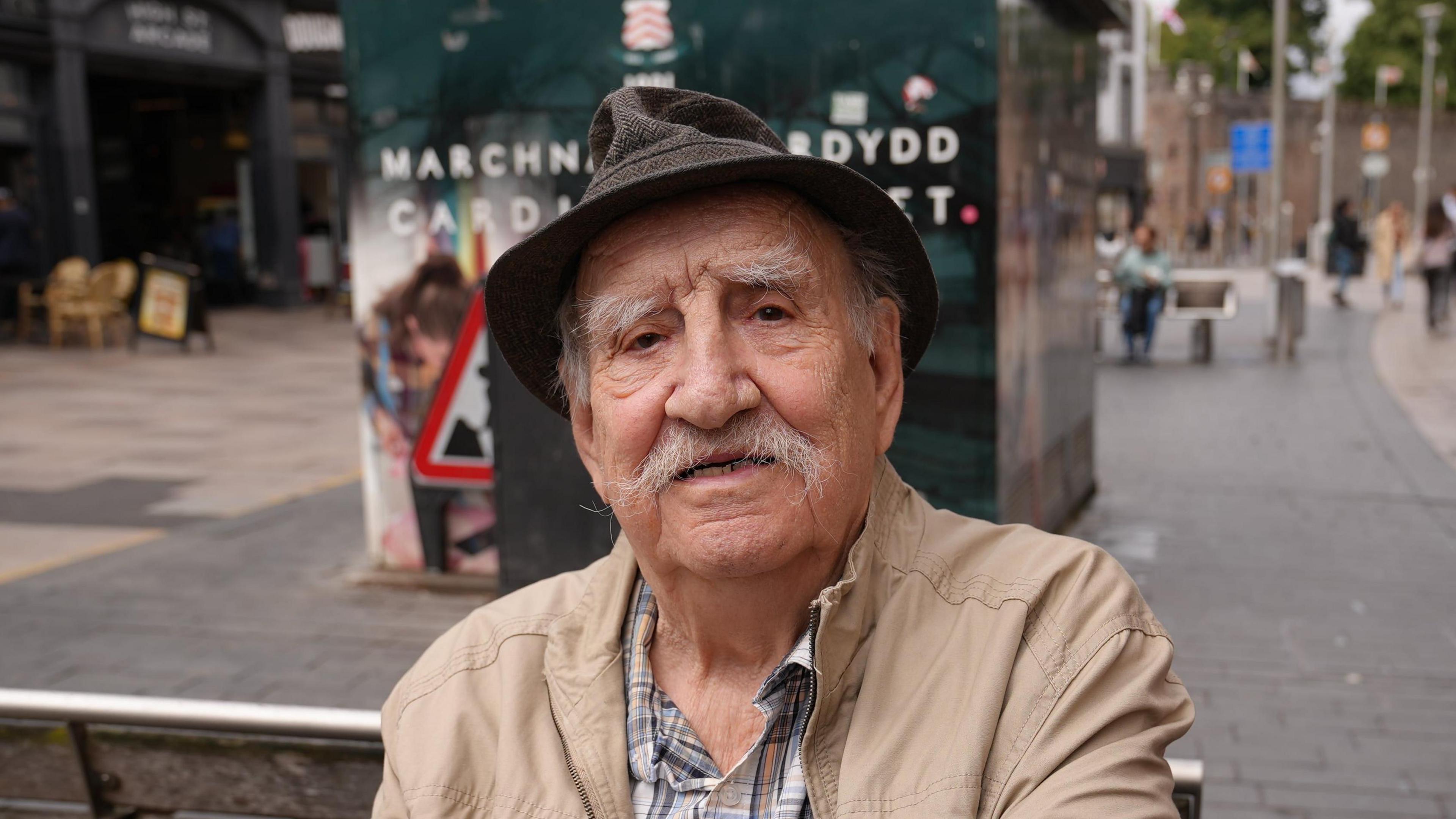 David Mort sat on a bench in Working Steet in Cardiff. He is wearing a hat and a beige raincoat and a checked shirt.