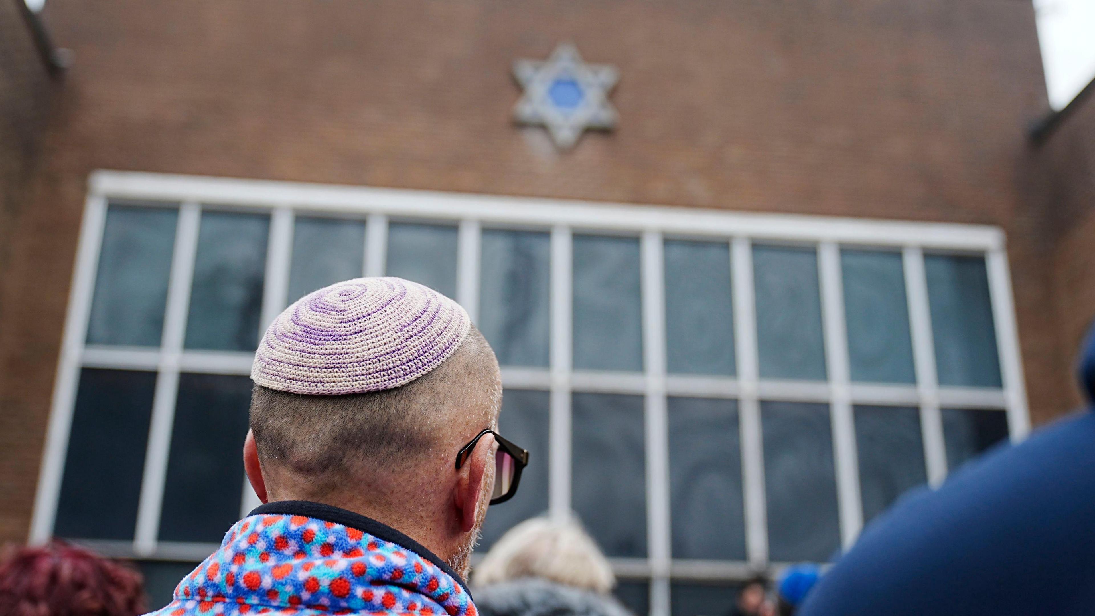 A man in a red, blue and purple spotted fleece and a Jewish Kippah cap stands as part of a crowd looking towards a brick building with a Star of David symbol mounted on the wall.