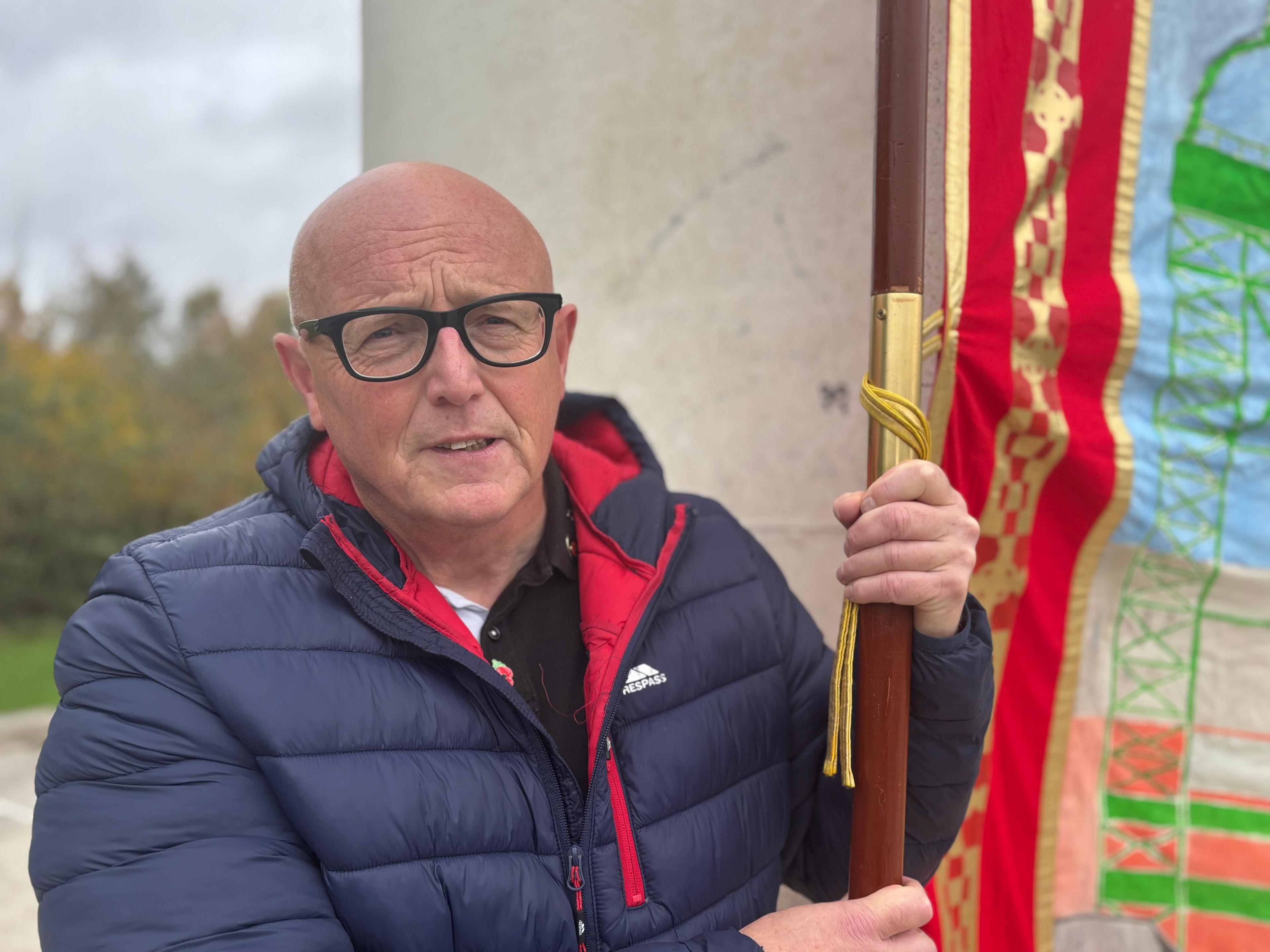 Jeff Bannister, who wears dark glasses and blue coat, poses as he holds the union banner in front of the sculpture.